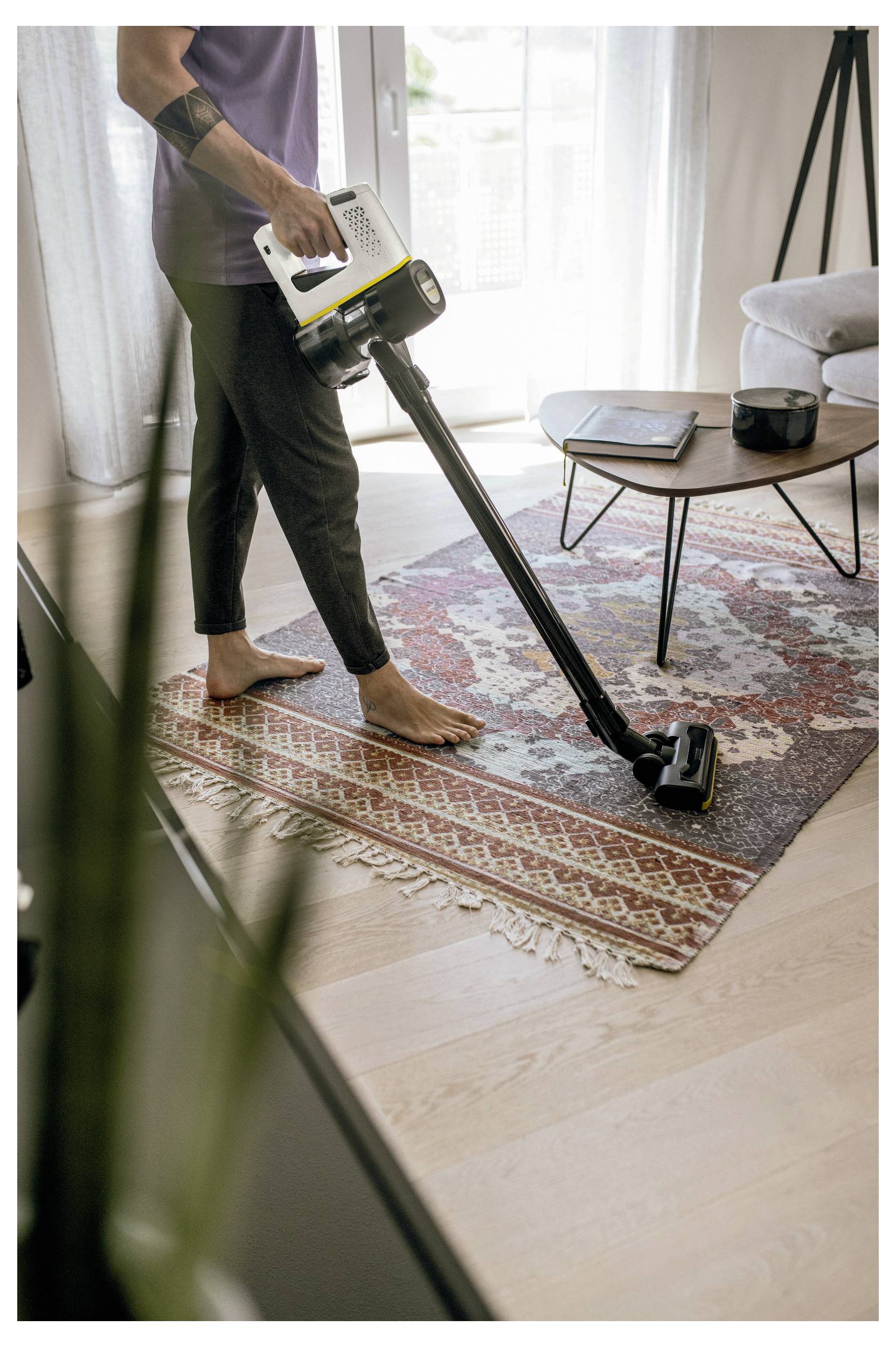 Eine Person saugt einen gemusterten Teppich in einem Wohnzimmer mit einem kabellosen Staubsauger. In der Nähe steht ein kleiner Tisch mit Gegenständen darauf.