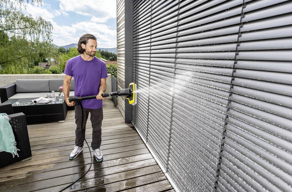 Ein Mann bedient einen Hochdruckreiniger an geschlossenen Fensterjalousien auf einer Holzterrasse und trägt ein lila Hemd und Jeans. Im Hintergrund ist Gartenmöbel zu sehen.