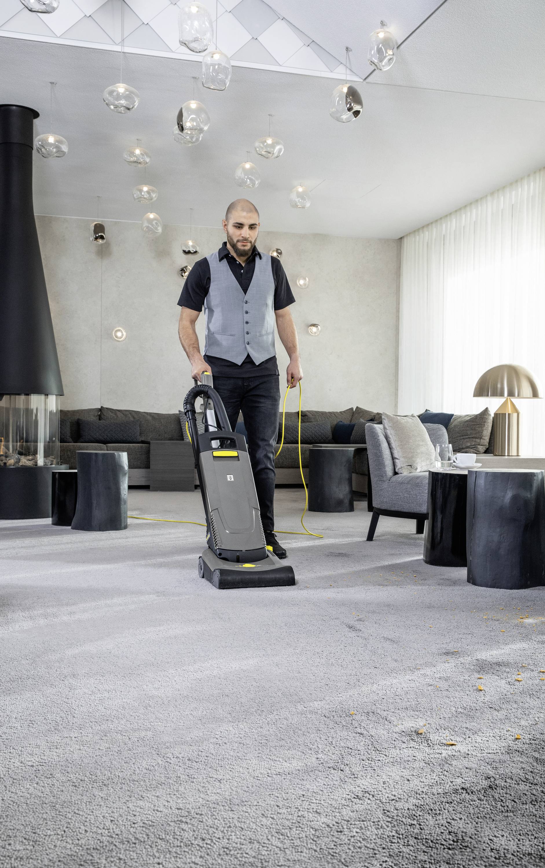 Eine Person saugt mit einem Staubsauger einen grauen Teppich in einem modern eingerichteten Wohnzimmer mit Kamin und Sofas.