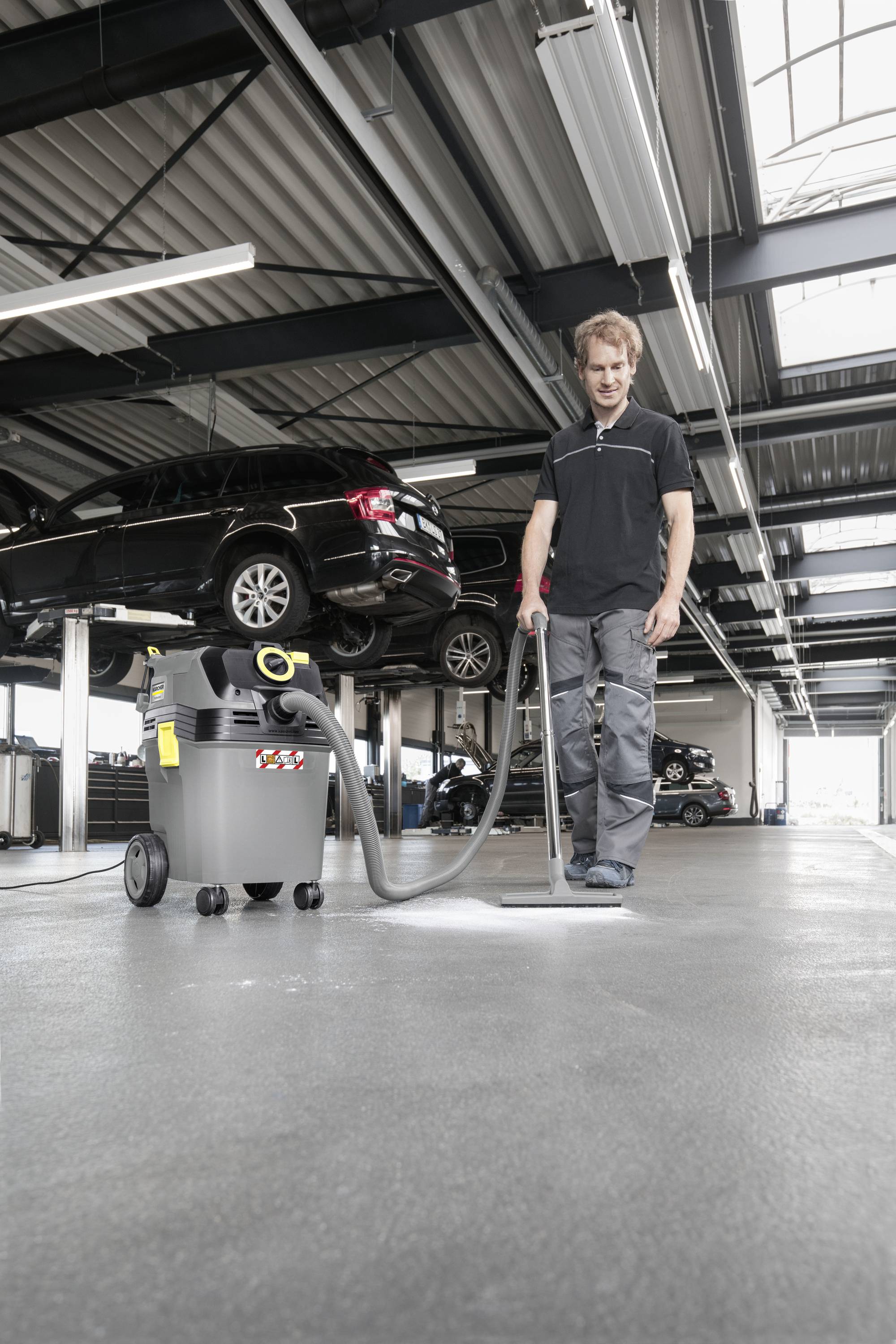 Ein Mechaniker reinigt den Boden einer Autowerkstatt mit einem Industriestaubsauger. Im Hintergrund sind Autos auf Hebebühnen.