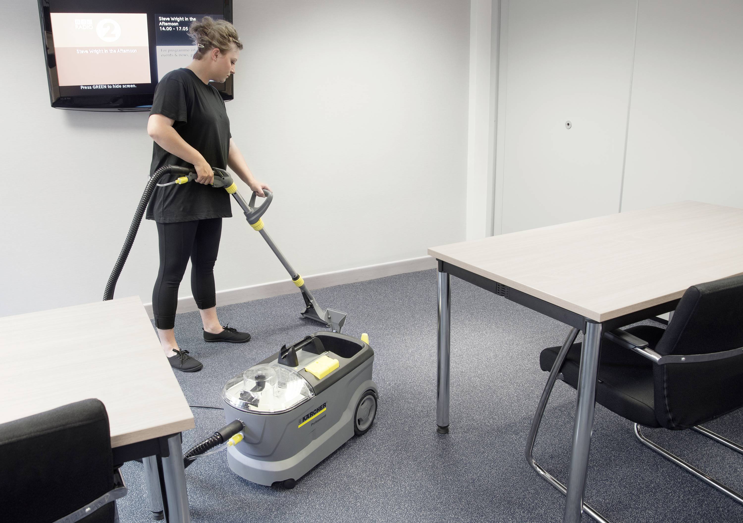Eine Person saugt mit einem Teppichreiniger den Boden in einem Konferenzraum mit grauem Teppich und zwei Tischen mit schwarzen Stühlen.