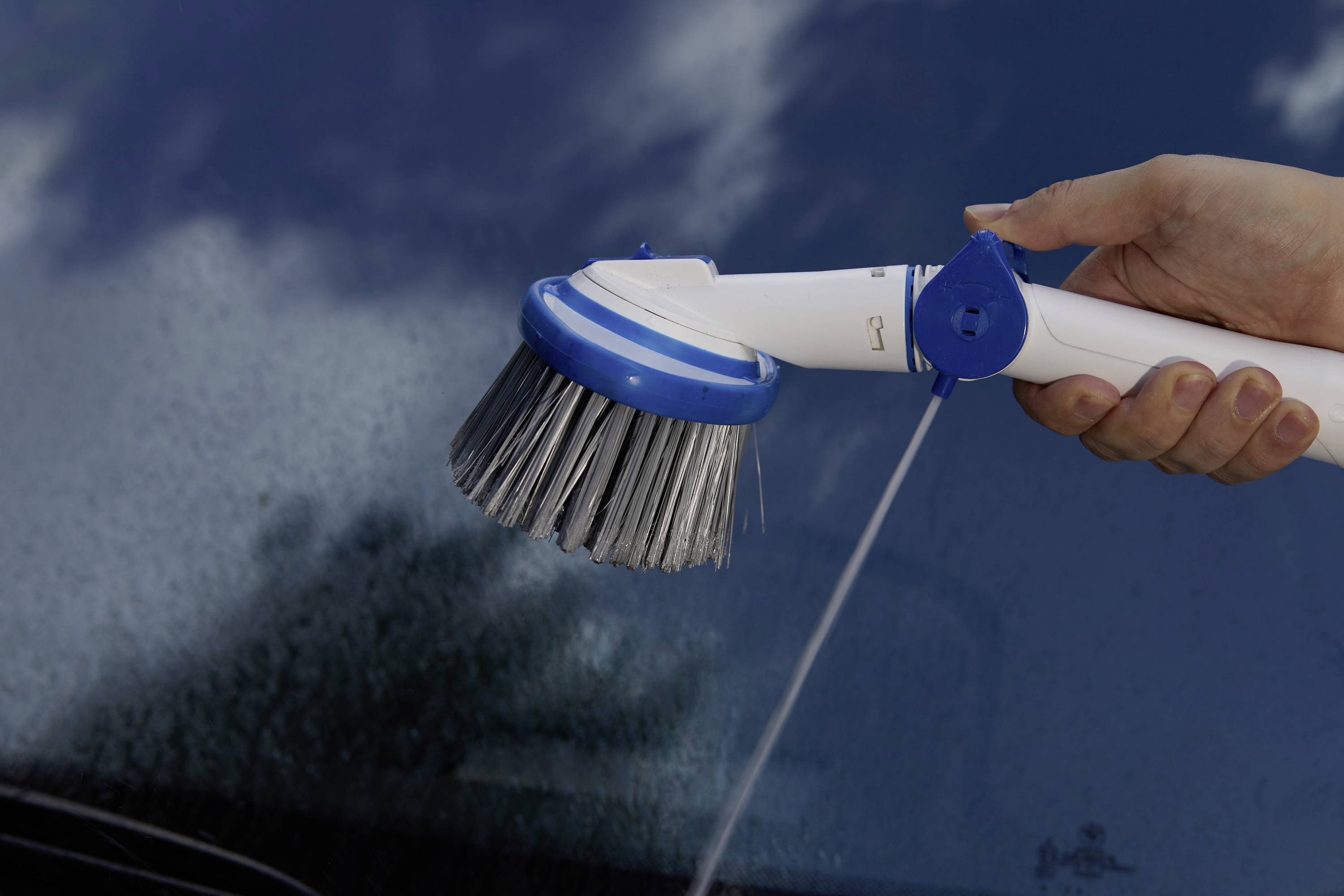 Eine Hand hält eine elektrische Reinigungsbürste mit Wasserstrahl über eine nasse Glasoberfläche, um diese zu säubern.