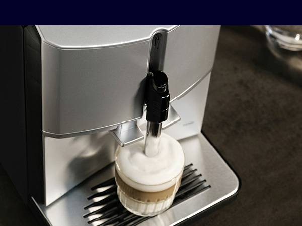 Eine Kaffeemaschine bereitet einen Cappuccino zu. Der Kaffee wird in eine Tasse gegossen, die auf einem silbernen Tropfgitter steht.