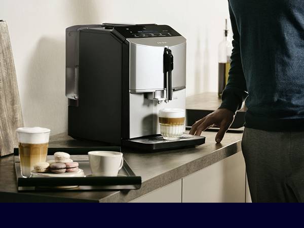 Eine Person bereitet einen Kaffee mit einem Kaffeevollautomaten zu. Auf dem Tisch stehen zwei Gläser mit Kaffee und einige Macarons.