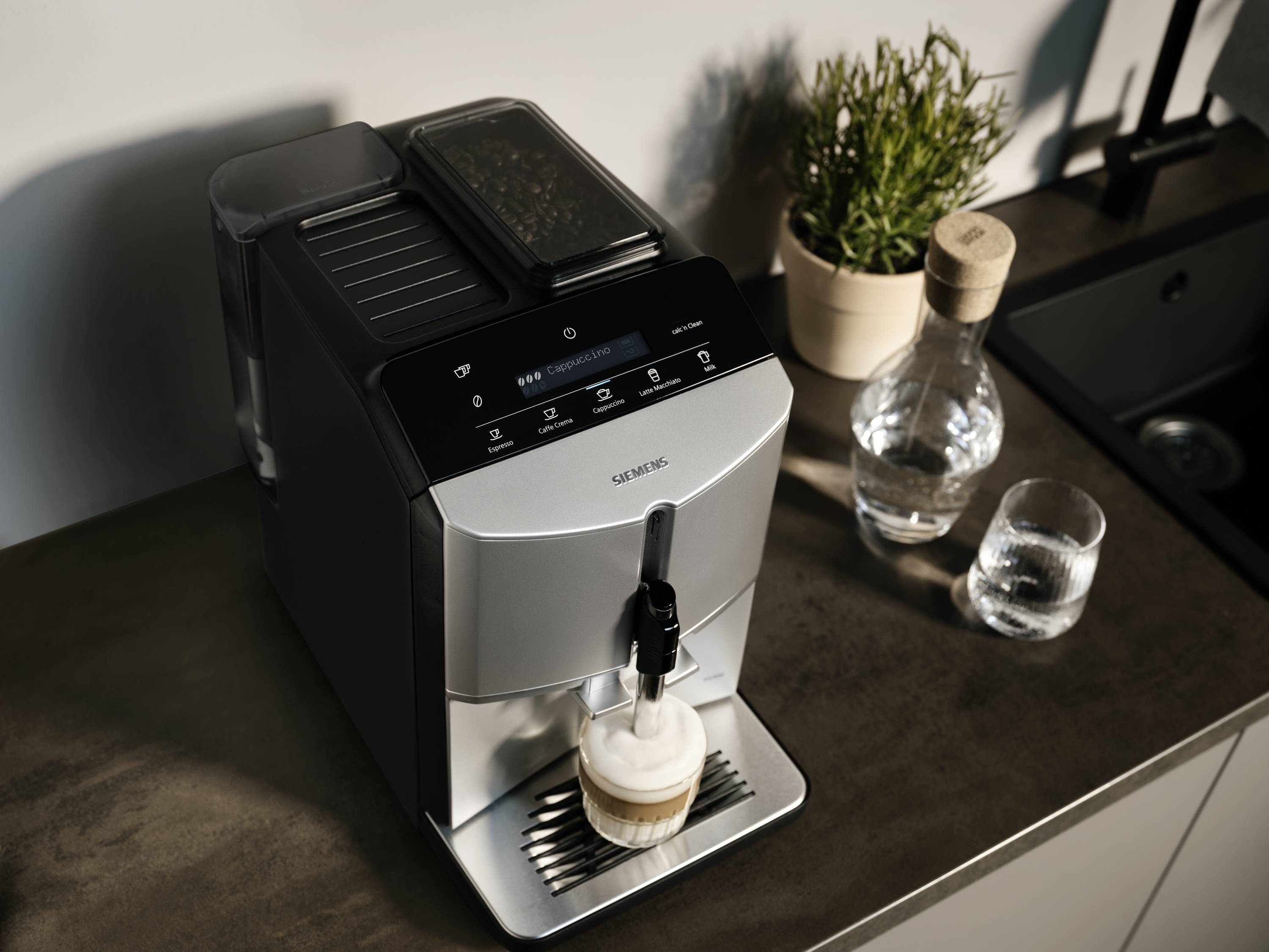 Kaffeevollautomat auf einer Küchentheke bereitet einen Latte Macchiato zu; daneben steht eine Karaffe Wasser und ein Glas.