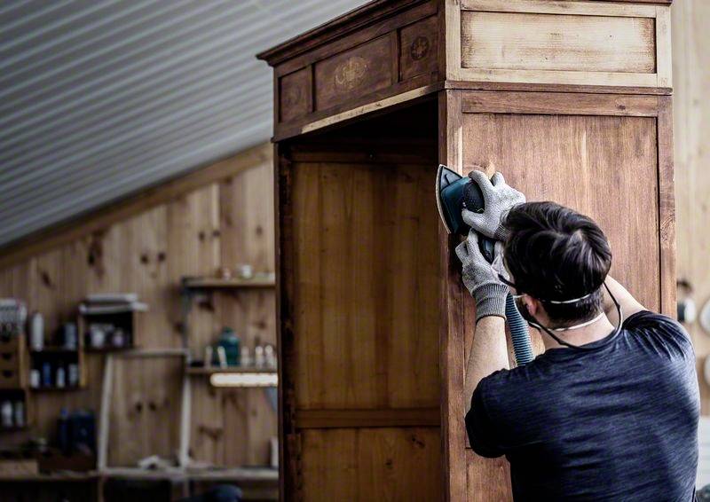 Ein Mann schleift einen hölzernen Schrank in einer Werkstatt. Er trägt Arbeitshandschuhe und ein Atemschutzgerät. Im Hintergrund sind Regale mit Werkzeugen sichtbar.