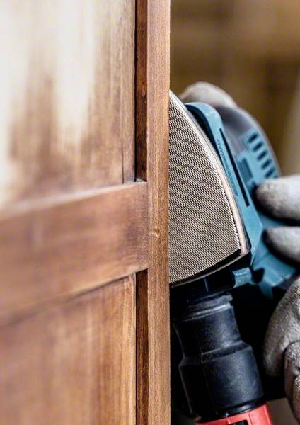 Eine Person bearbeitet die Kante eines Holzmöbels mit einem elektrischen Schleifgerät. Die Szene zeigt handwerkliche Tätigkeit.