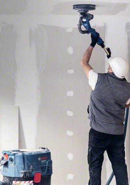 Ein Bauarbeiter mit Schutzhelm und Gerät schleift eine Wand. Im Hintergrund sind unfertige Wände und Werkzeuge zu sehen.