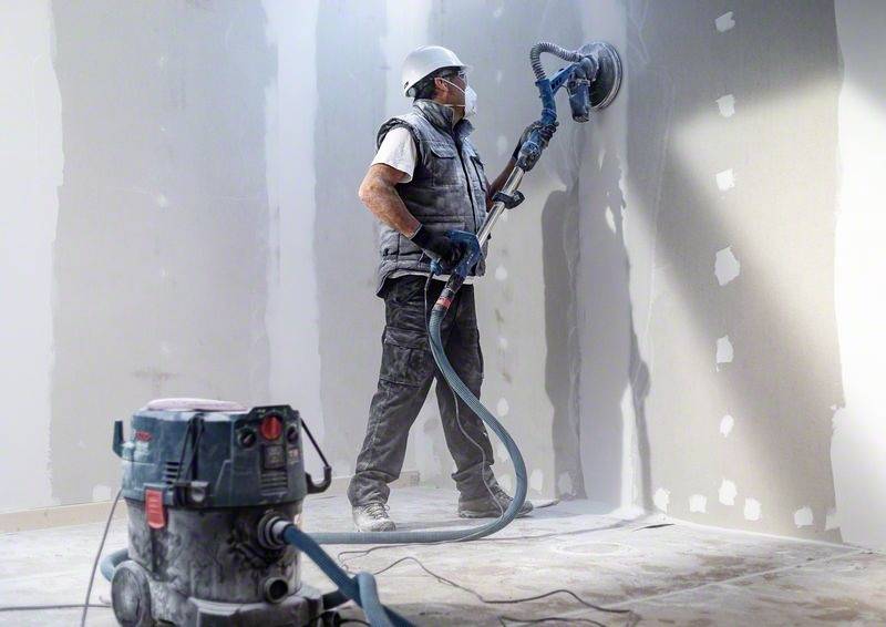 Ein Arbeiter mit Schutzhelm und Maske glättet mit einer Schleifmaschine eine Wand in einem Raum. Neben ihm steht ein Staubsauger.