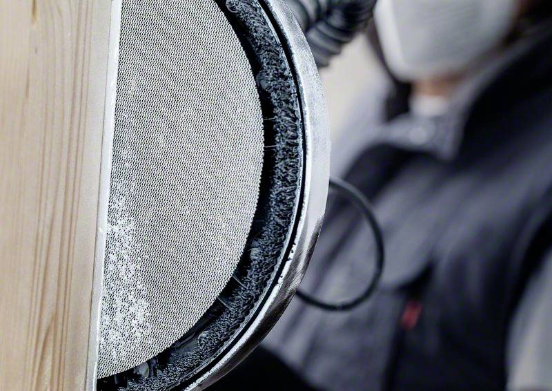 Ein Mensch schleift eine Holzoberfläche mit einem elektrischen Schleifgerät. Eine Staubmaske wird zum Schutz getragen.