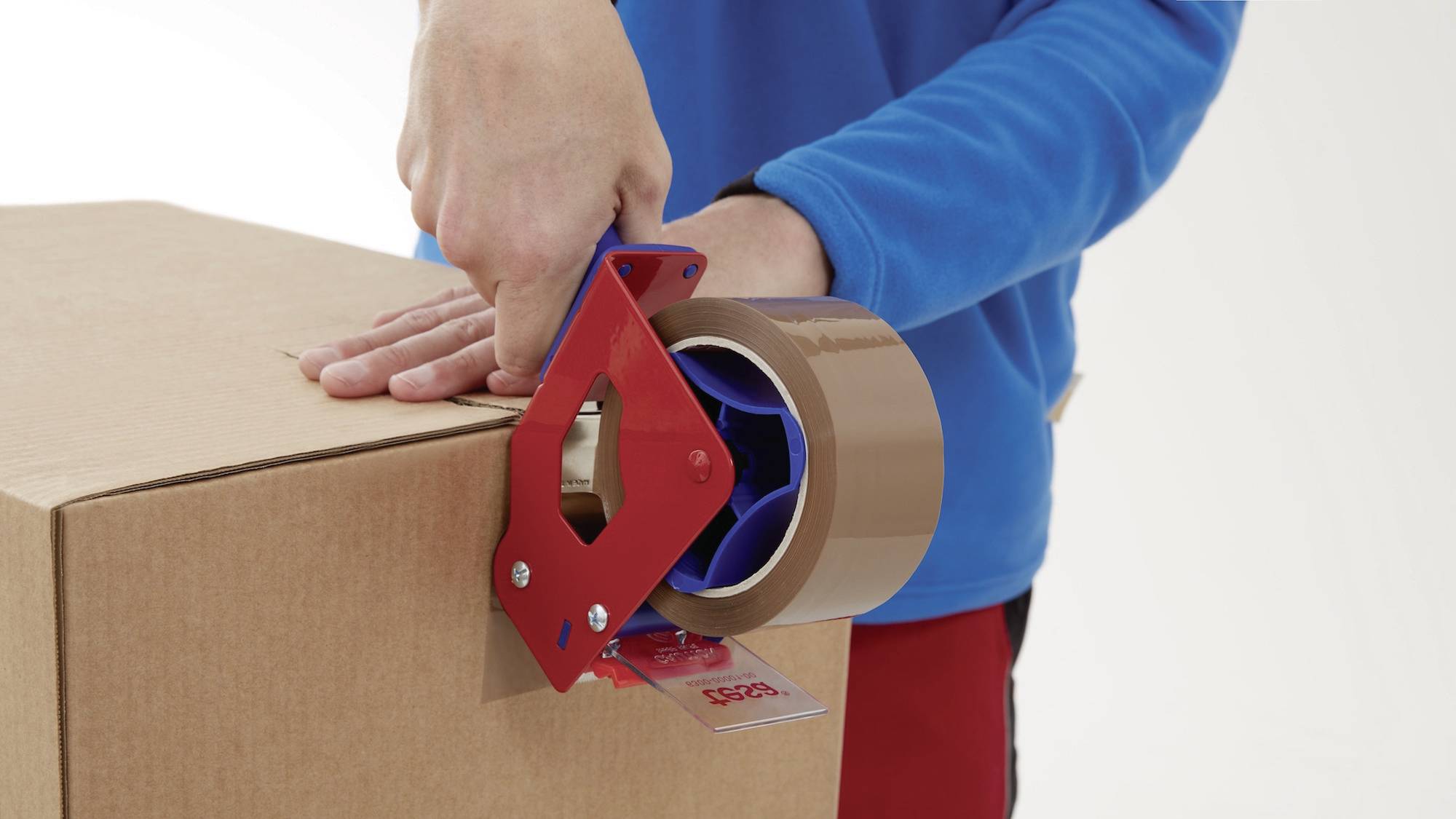 Eine Person verschließt einen Karton mit braunem Klebeband, das in einem roten Hand-Abroller gehalten wird.