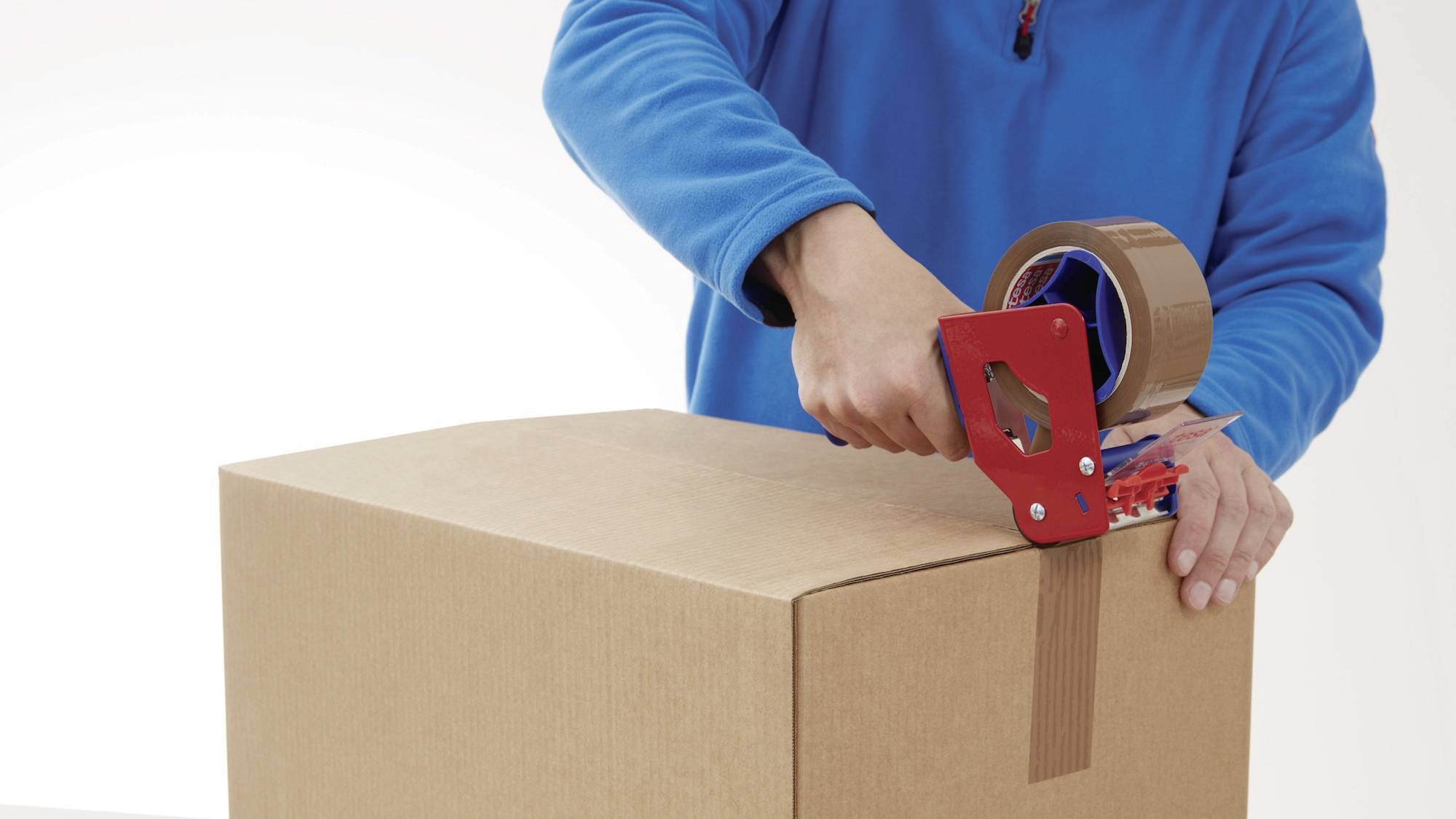 Eine Person klebt mit einem roten Handabroller braunes Klebeband auf einen Karton, um ihn zu verschließen. Sie trägt eine blaue Jacke.
