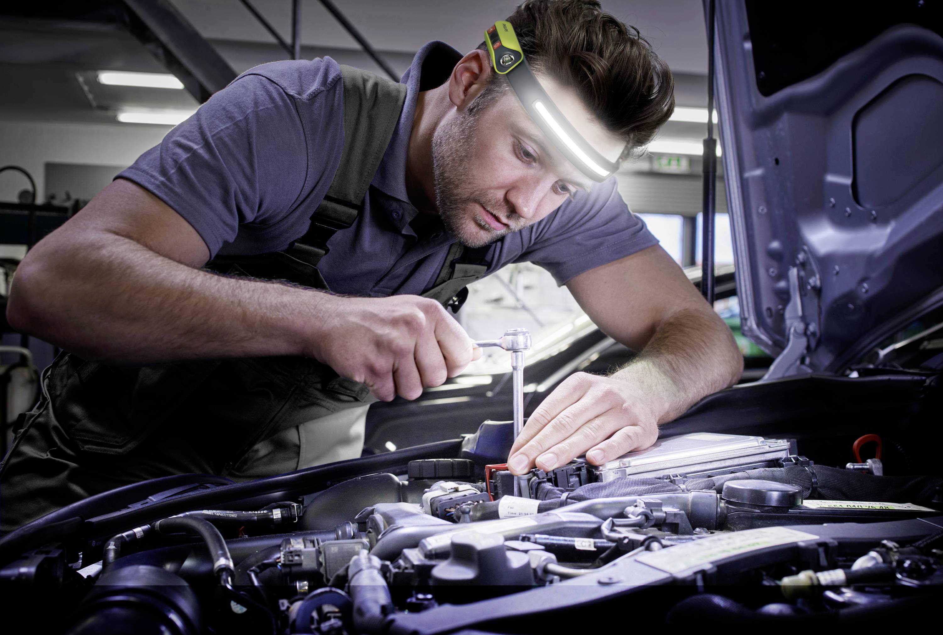 Ein Mechaniker, der konzentriert an einem Auto-Motor arbeitet, trägt eine Stirnlampe. Im Hintergrund sind Werkzeuge und Werkstattbeleuchtung sichtbar.
