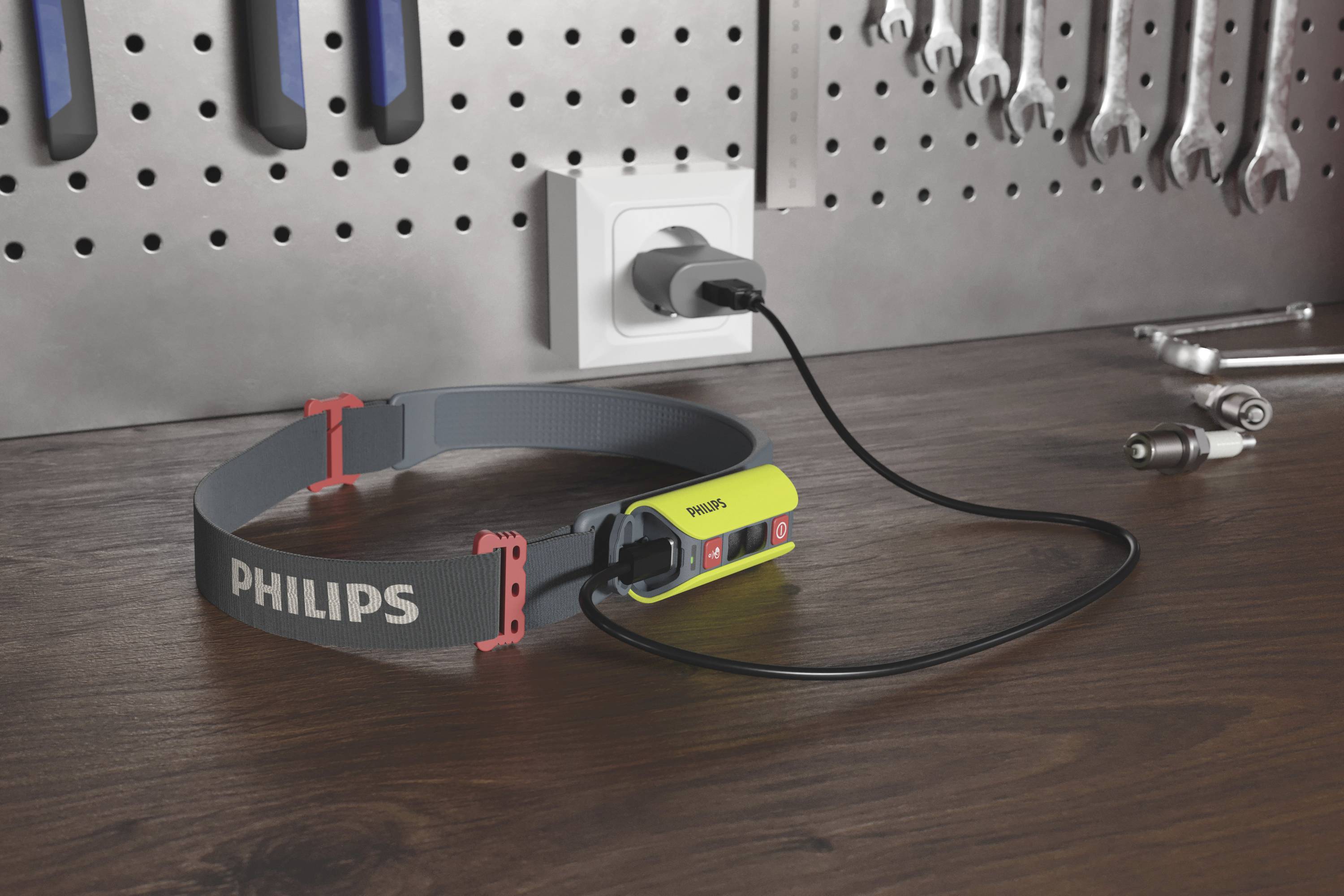 Eine Stirnlampe der Marke Philips liegt auf einer Werkbank, angeschlossen an eine Steckdose. Im Hintergrund hängen Werkzeuge an einer Wand.