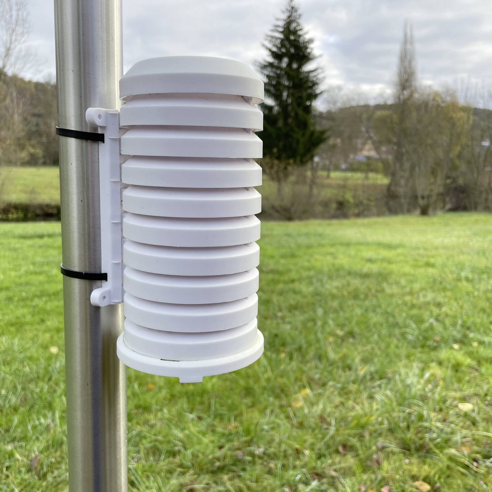 Ein weißer Wettersensor an einem Metallpfosten misst klimatische Daten. Im Hintergrund sind eine Wiese und Bäume sichtbar.