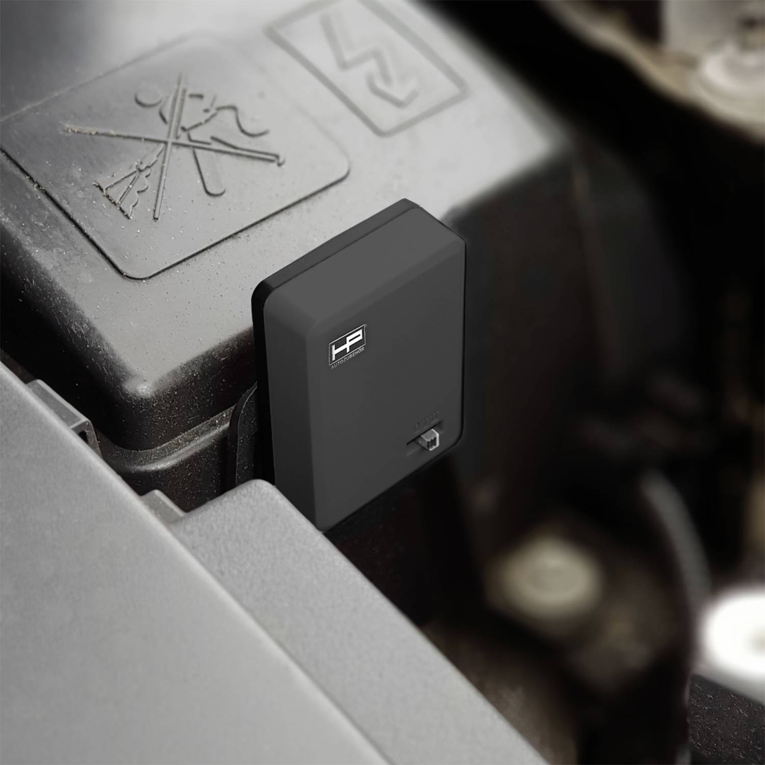 Ein kleines schwarzes Gerät mit einem Logo ist unter der Motorhaube eines Fahrzeugs installiert, das teilweise eine geprägte Warnaufkleber verdeckt.