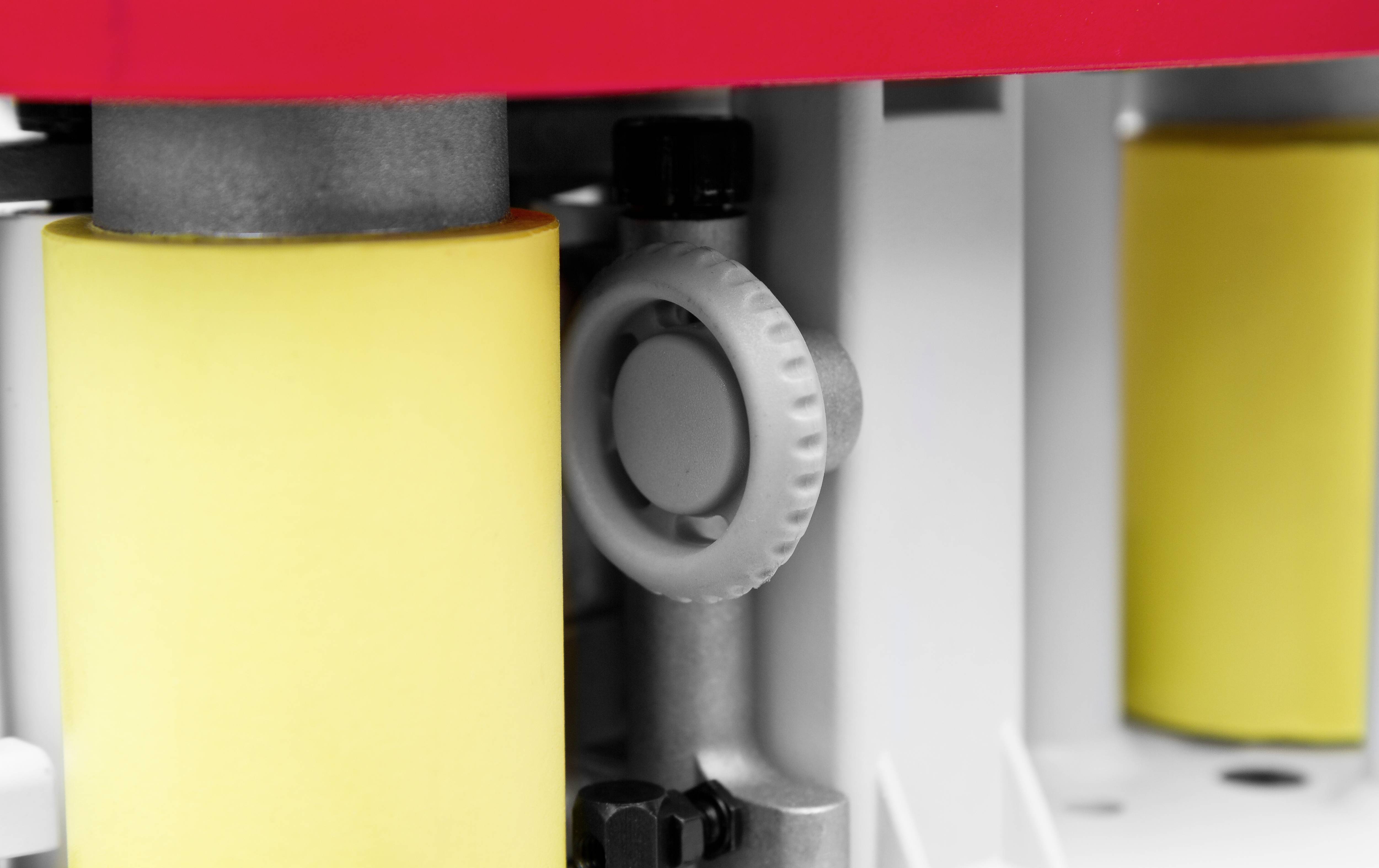 Maschinenteil mit gelben Rollen und einem grauen Drehknopf, teilweise verdeckt. Technische Umgebung, rote Abdeckung oben.