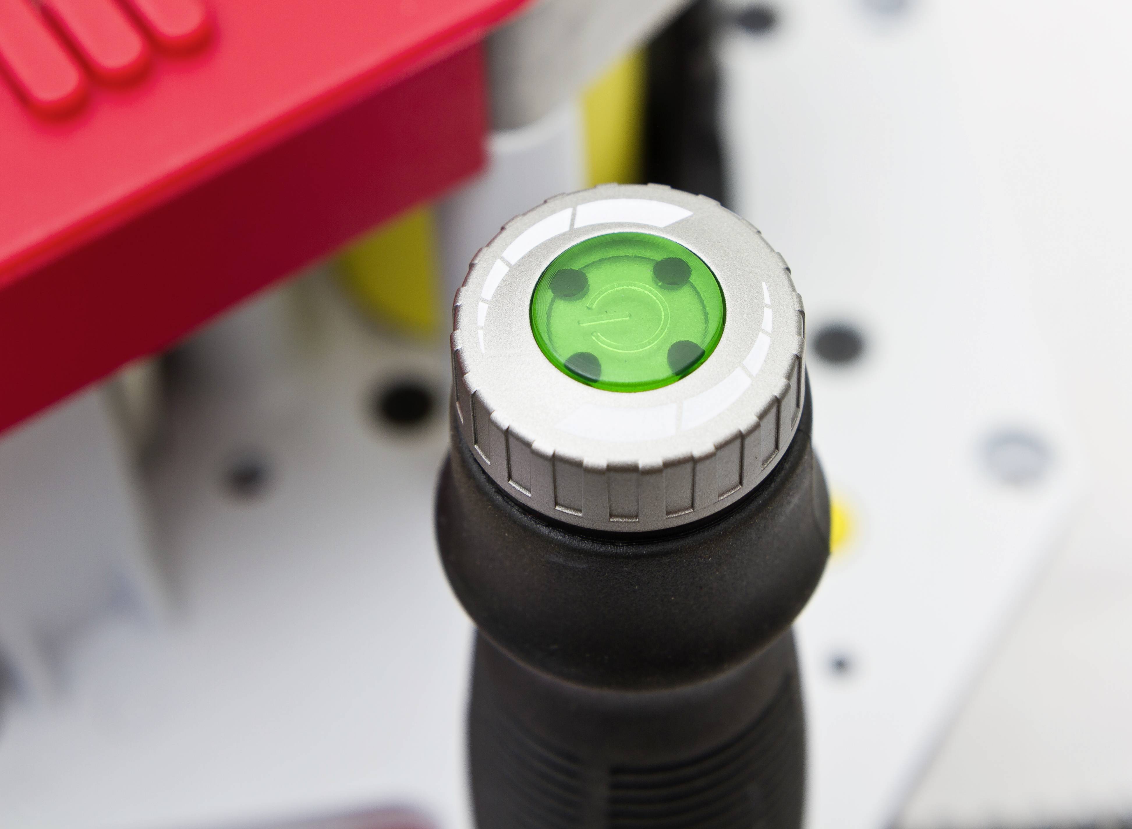 Griff eines Werkzeugs mit einem grünen Kontrollknopf, vermutlich zur Bedienung oder Kontrolle der Maschine im Hintergrund.