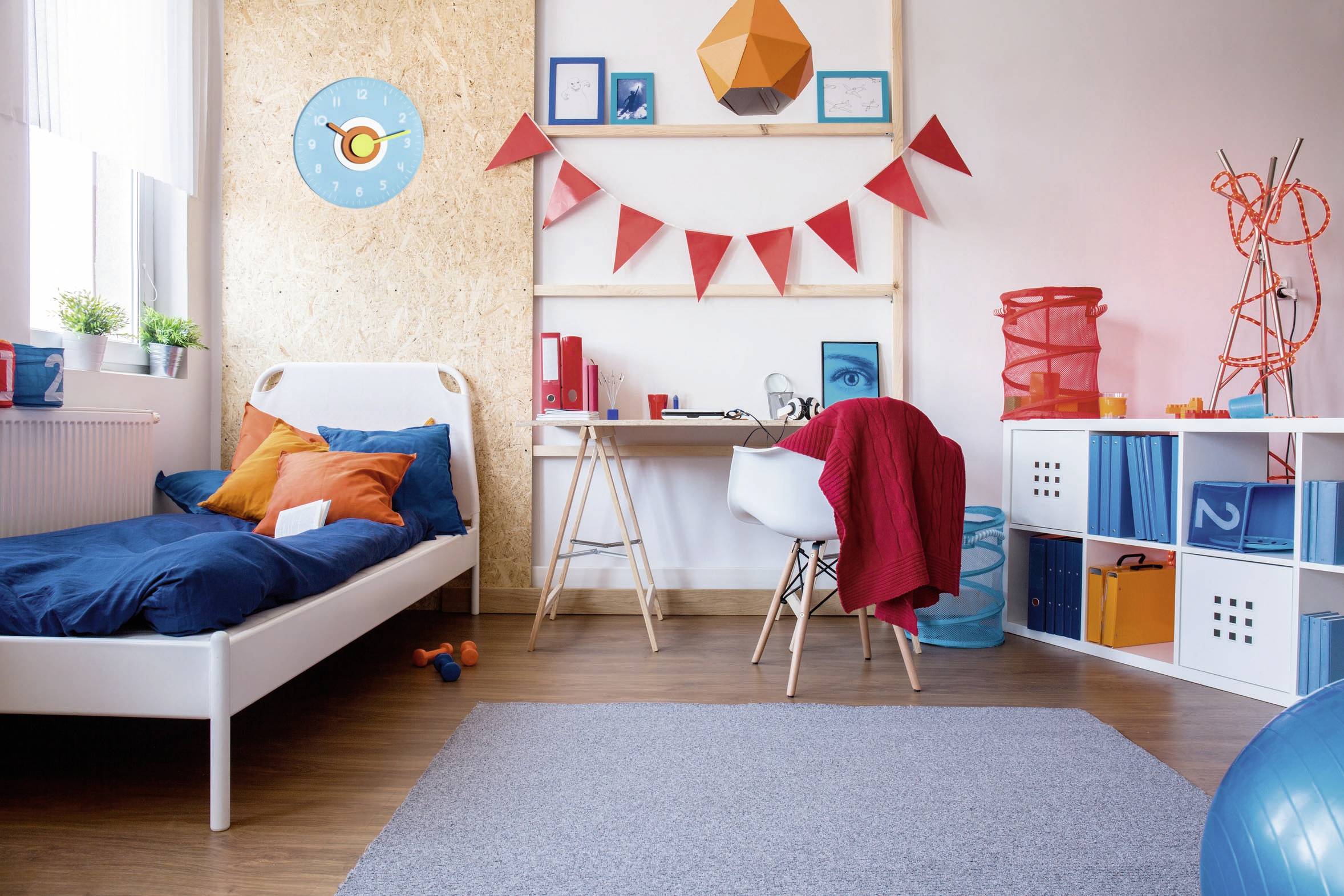 Ein modernes Kinderzimmer mit blauen und orangefarbenen Akzenten. Links ein Bett, rechts ein Schreibtisch, darüber bunte Wimpel.
