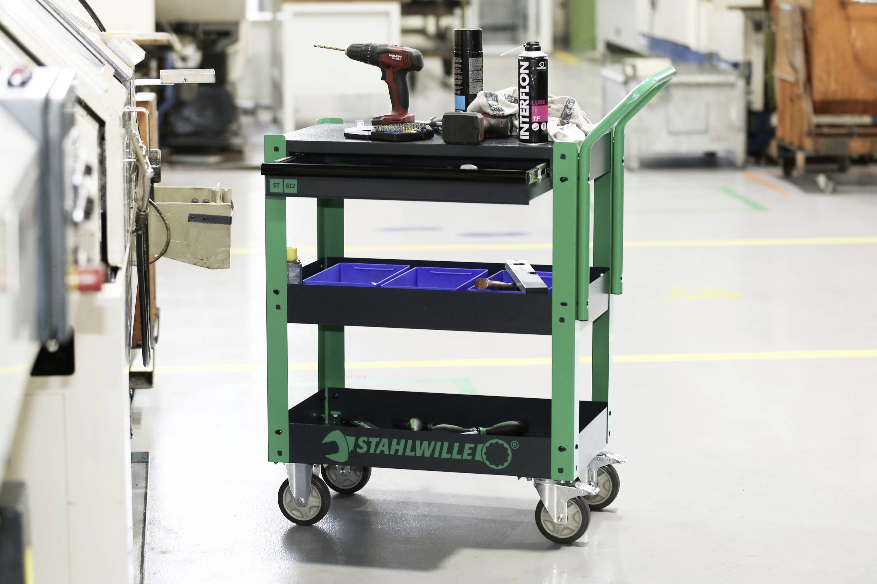 Ein grüner Werkzeugwagen mit drei Ablageflächen steht in einer Werkstatt. Auf dem oberen Fach befinden sich ein Schraubenzieher und Werkzeugteile.