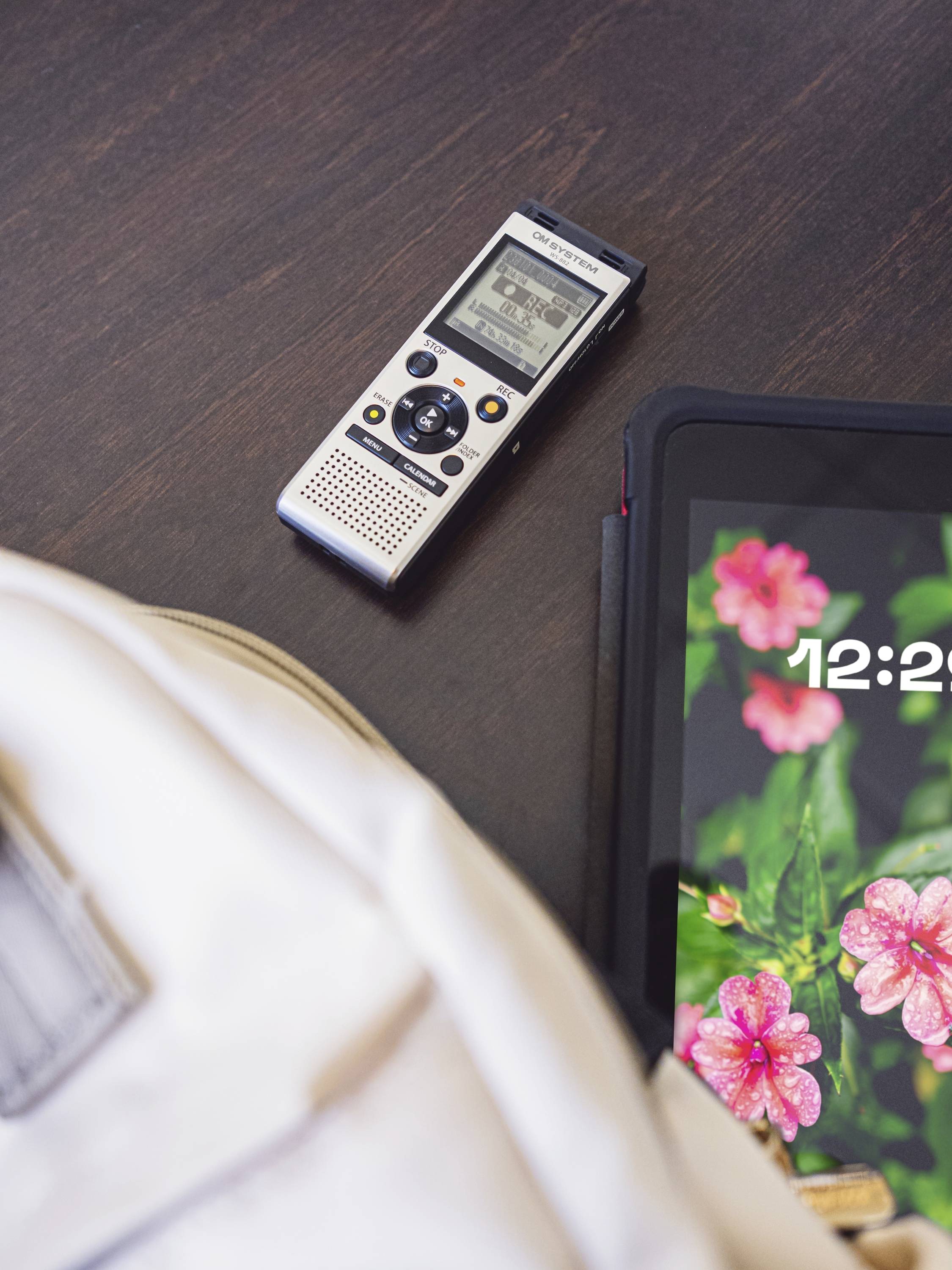 Ein schwarzer Tisch mit einem digitalen Diktiergerät und einem Tablet. Das Tablet zeigt die Uhrzeit '12:22' und ein Blumenmotiv.