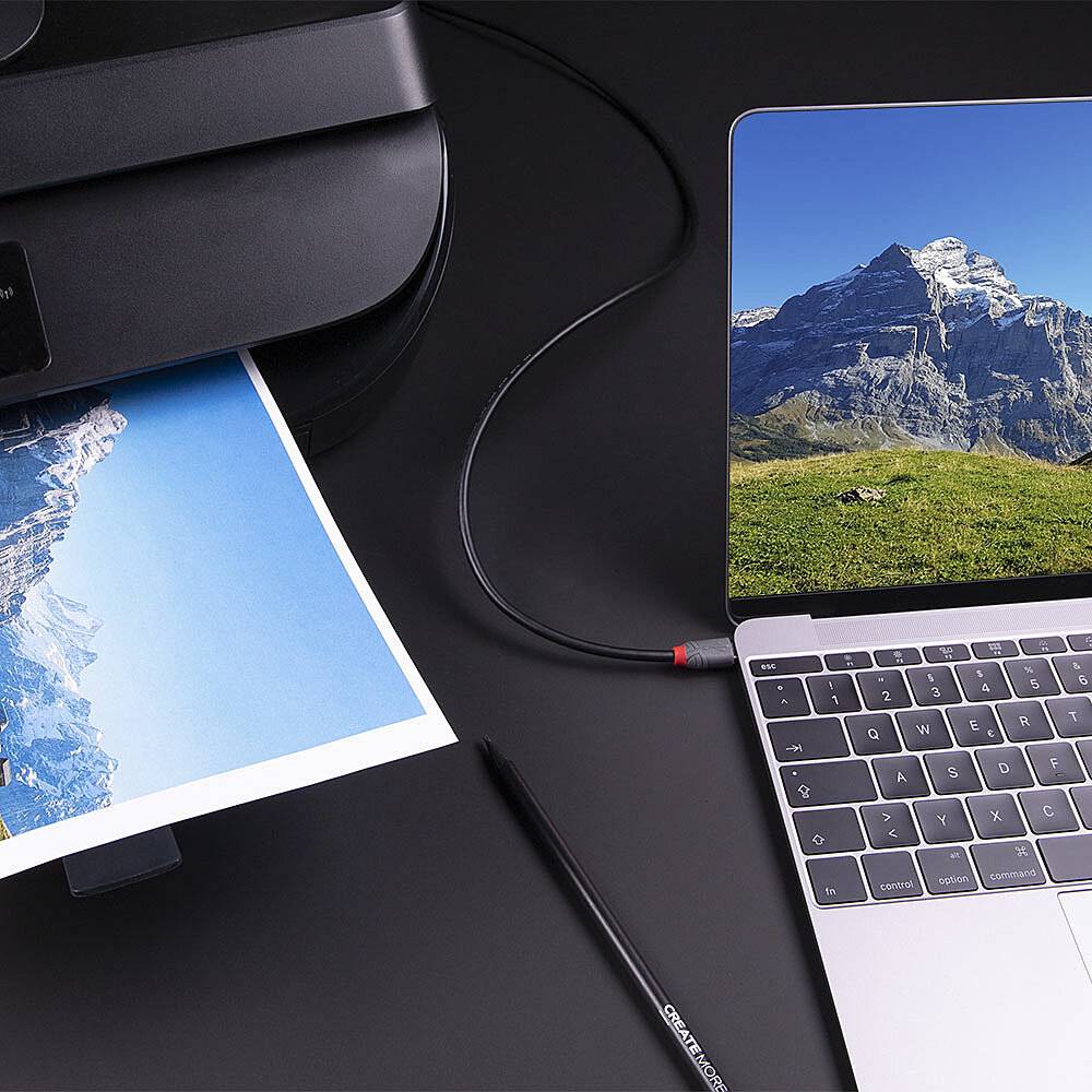 Drucker auf Schreibtisch druckt ein Foto der Berge, Laptop zeigt dasselbe Bild auf dem Bildschirm. Kabel verbindet Drucker und Laptop.