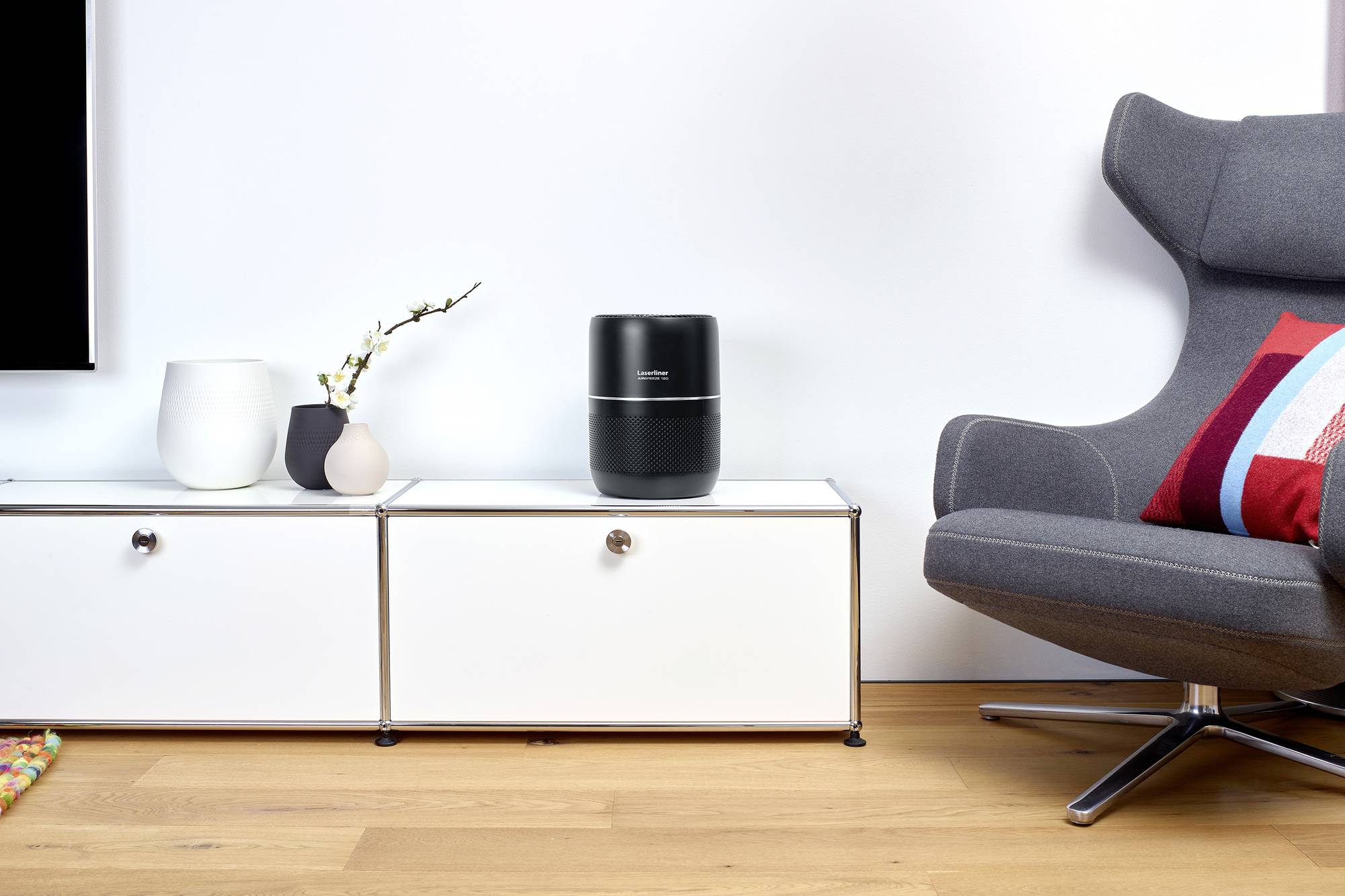 Ein moderner Wohnraum mit einem schwarzen Lautsprecher auf einem weißen Sideboard. Daneben stehen Vasen mit einer Orchidee. Rechts ein grauer Sessel mit buntem Kissen.