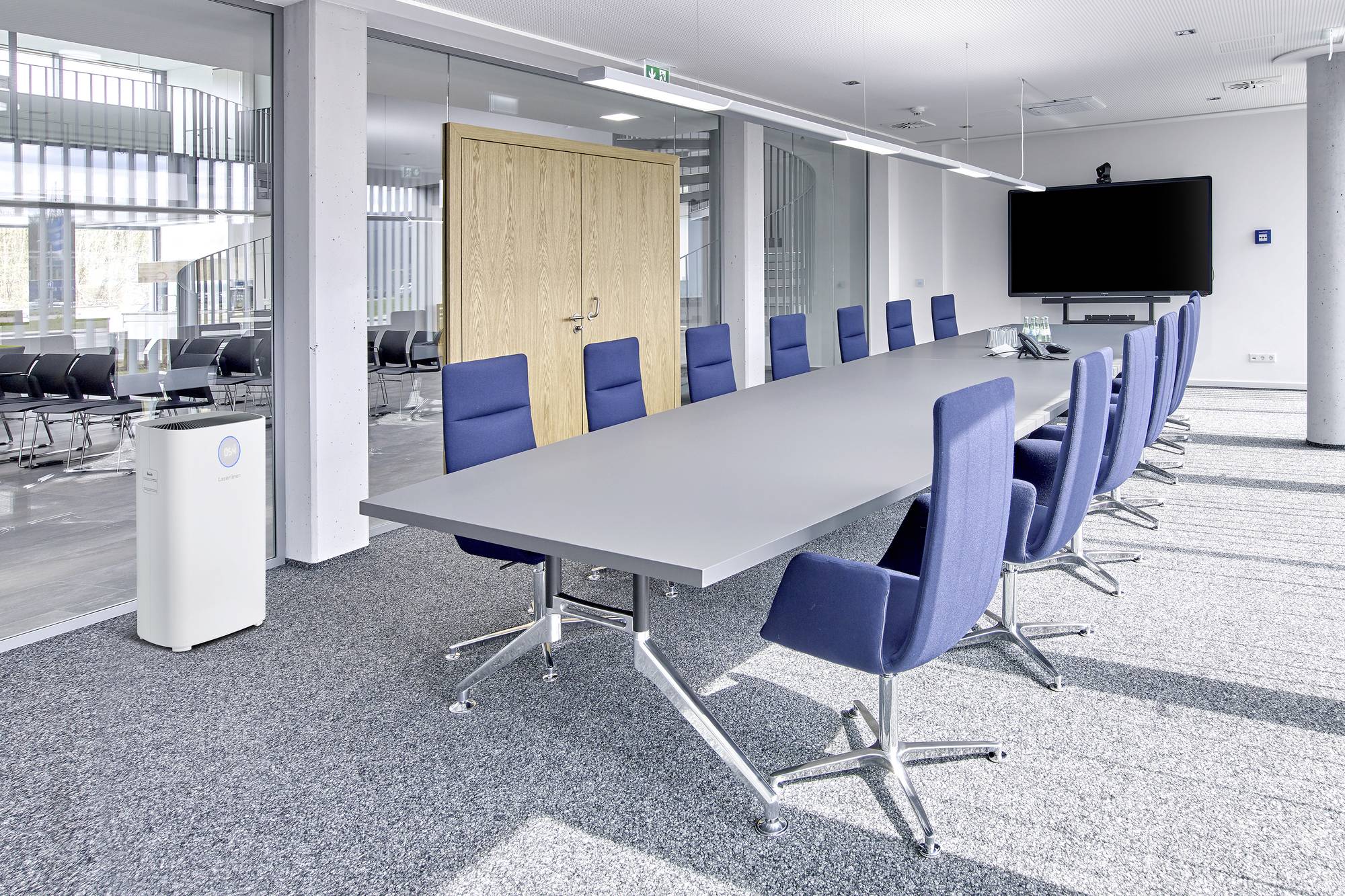 Konferenzraum mit großem rechteckigem Tisch und blauen Stühlen. An der Wand hängt ein großer Bildschirm. Raum ist hell und modern.