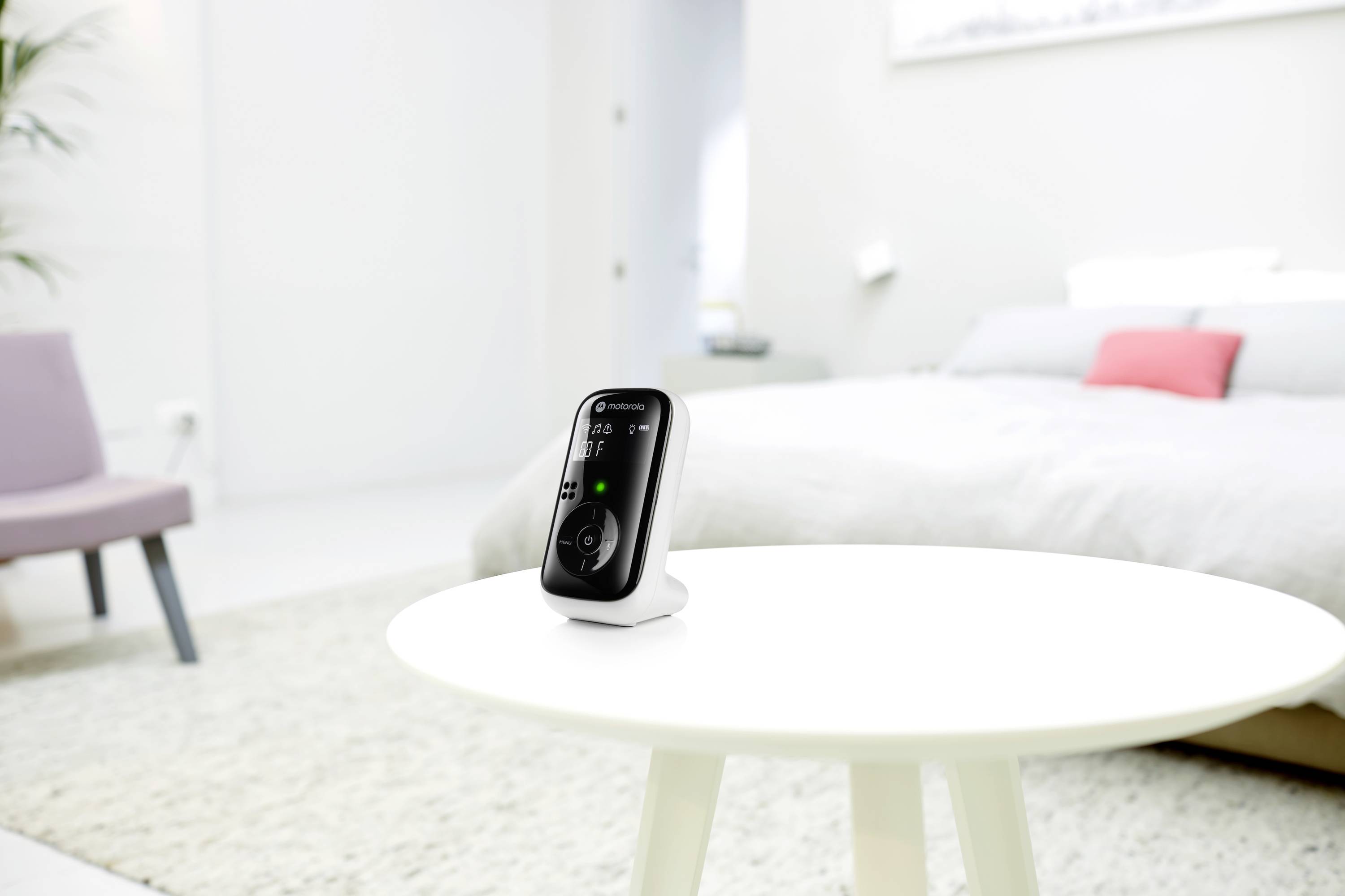 Ein Babymonitor steht eingeschaltet auf einem weißen Tisch in einem hellen Schlafzimmer. Im Hintergrund ist ein Bett zu sehen.