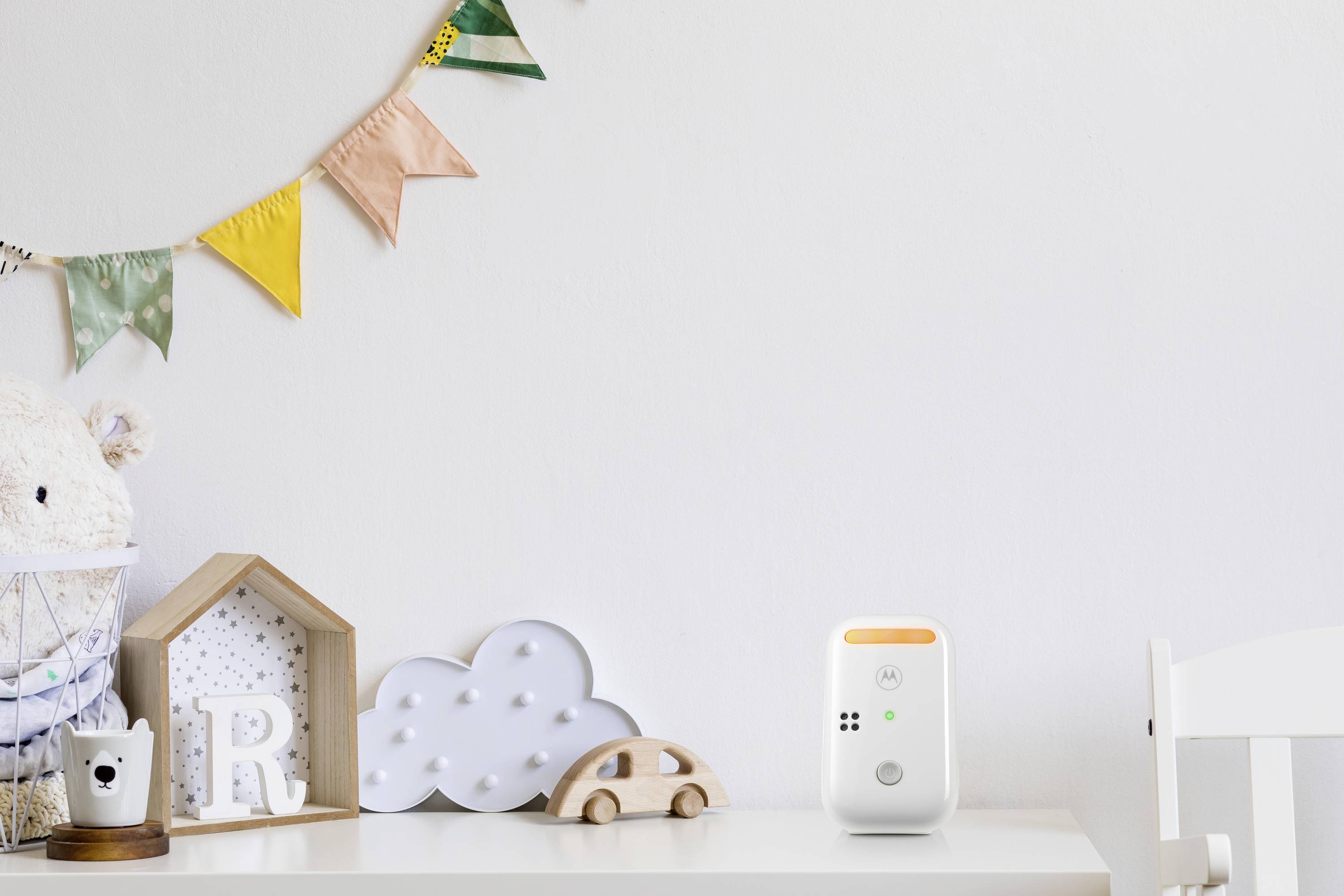 Ein Babyphone steht auf einem Tisch in einem Kinderzimmer. Im Hintergrund sind bunte Wimpel und Spielzeugdekorationen zu sehen.