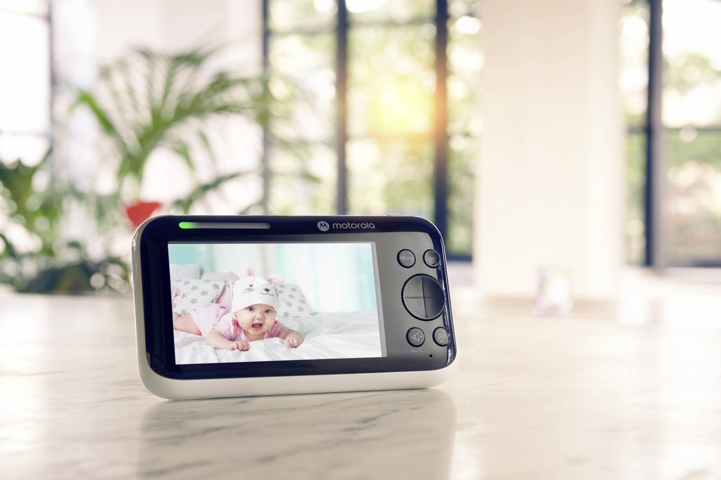 Ein Babyphone liegt auf einem Tisch, zeigt ein Video von einem lächelnden Baby. Im Hintergrund sind eine Pflanze und große Fenster.