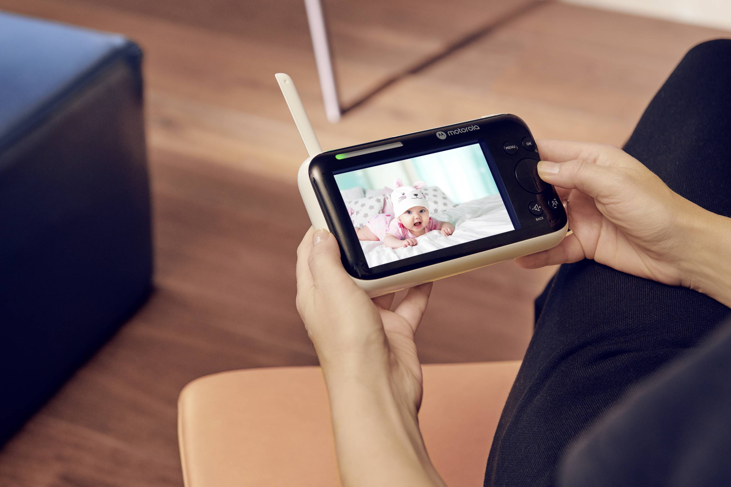 Eine Person hält ein Babyphon mit Videobildschirm in den Händen. Auf dem Bildschirm ist ein Baby mit Mütze zu sehen, das auf einer Decke liegt.