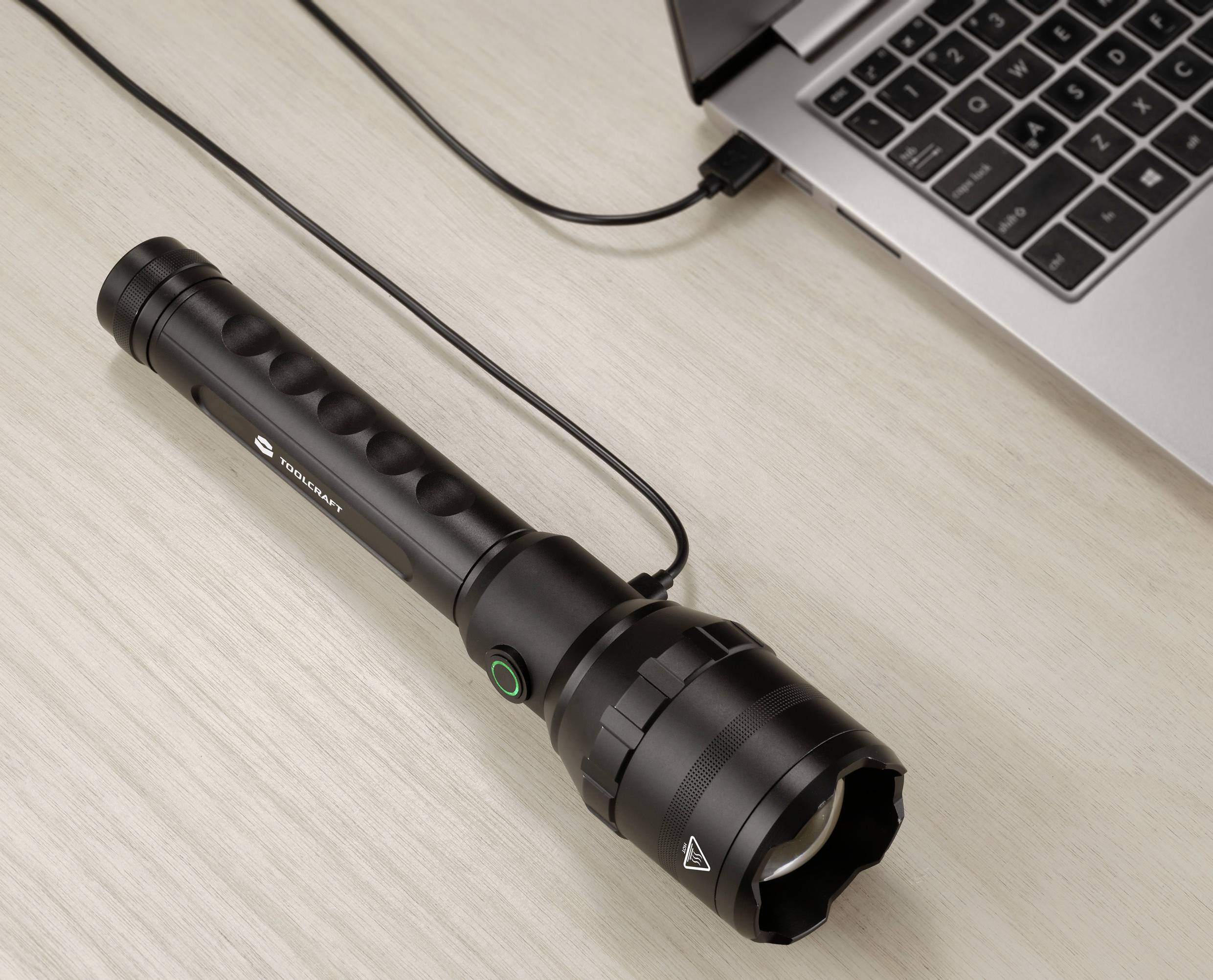 Eine schwarze Taschenlampe wird über ein USB-Kabel mit einem Laptop verbunden, auf einem Holztisch liegend.