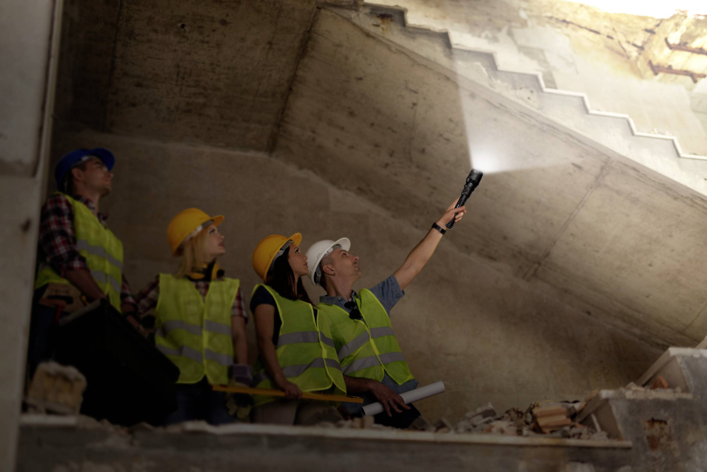 Arbeiter inspizieren eine Baustelle in einem Rohbau. Eine Person leuchtet mit einer Taschenlampe an die Decke. Alle tragen Schutzhelme und Warnwesten.