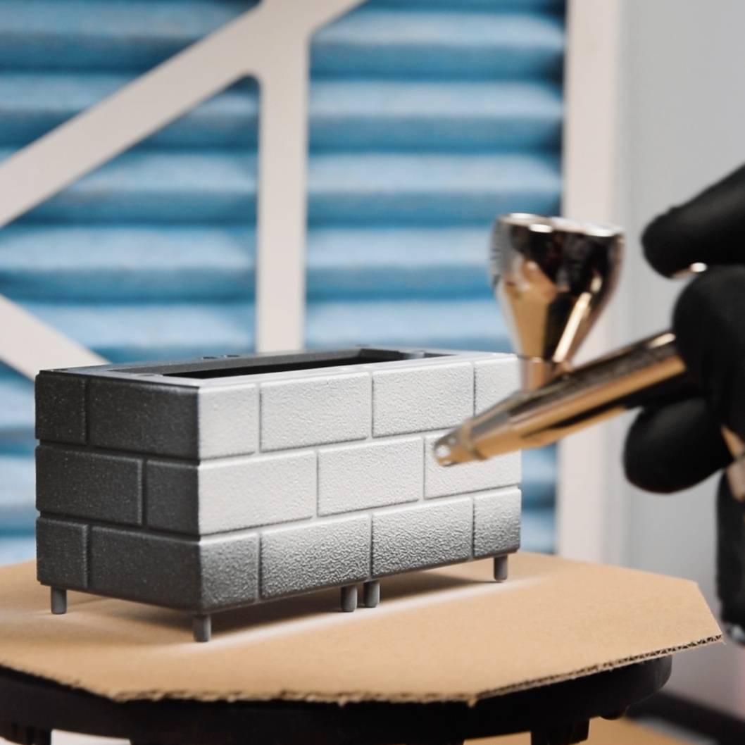 Eine Person sprüht mit einer Airbrush-Farbe auf ein graues, steinähnliches Modell, möglicherweise ein Miniaturbauwerk.