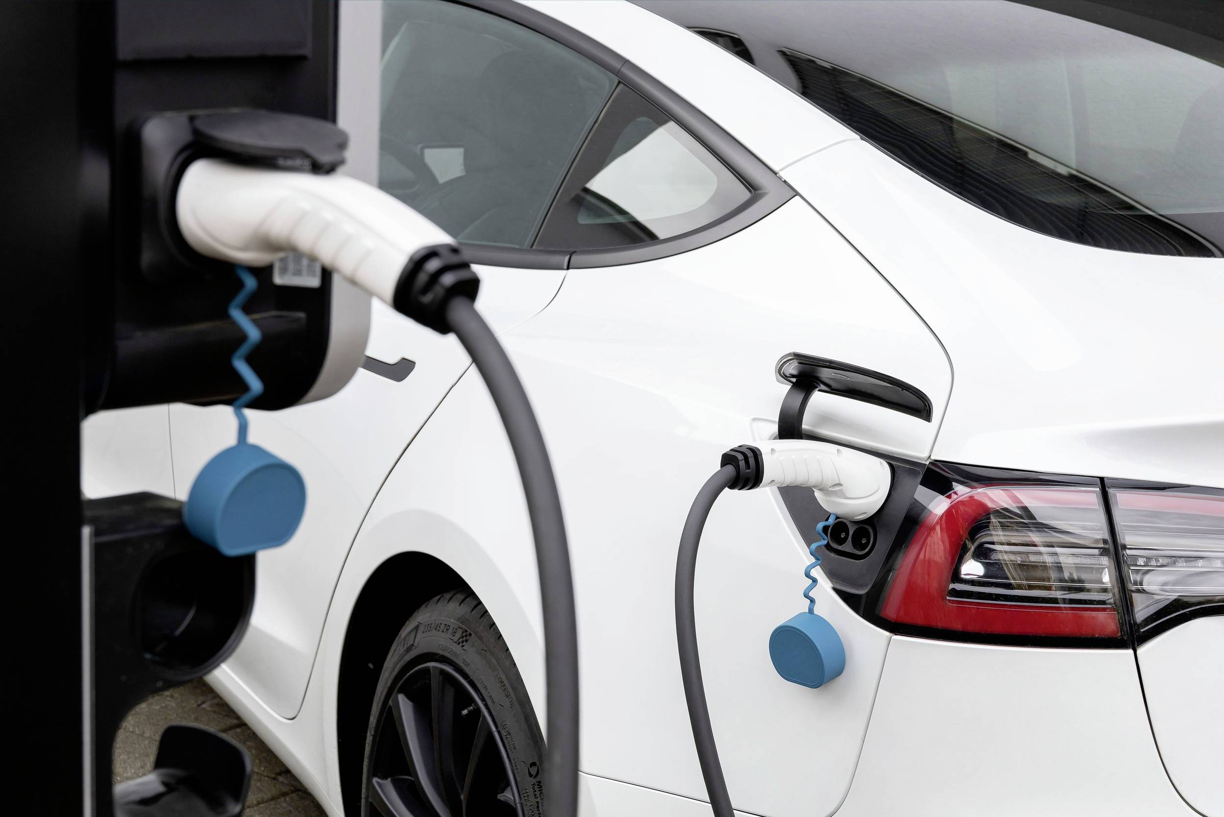 Weißes Elektroauto, das an einer Ladestation angeschlossen ist. Ladekabel sind an der Rückseite des Fahrzeugs montiert.