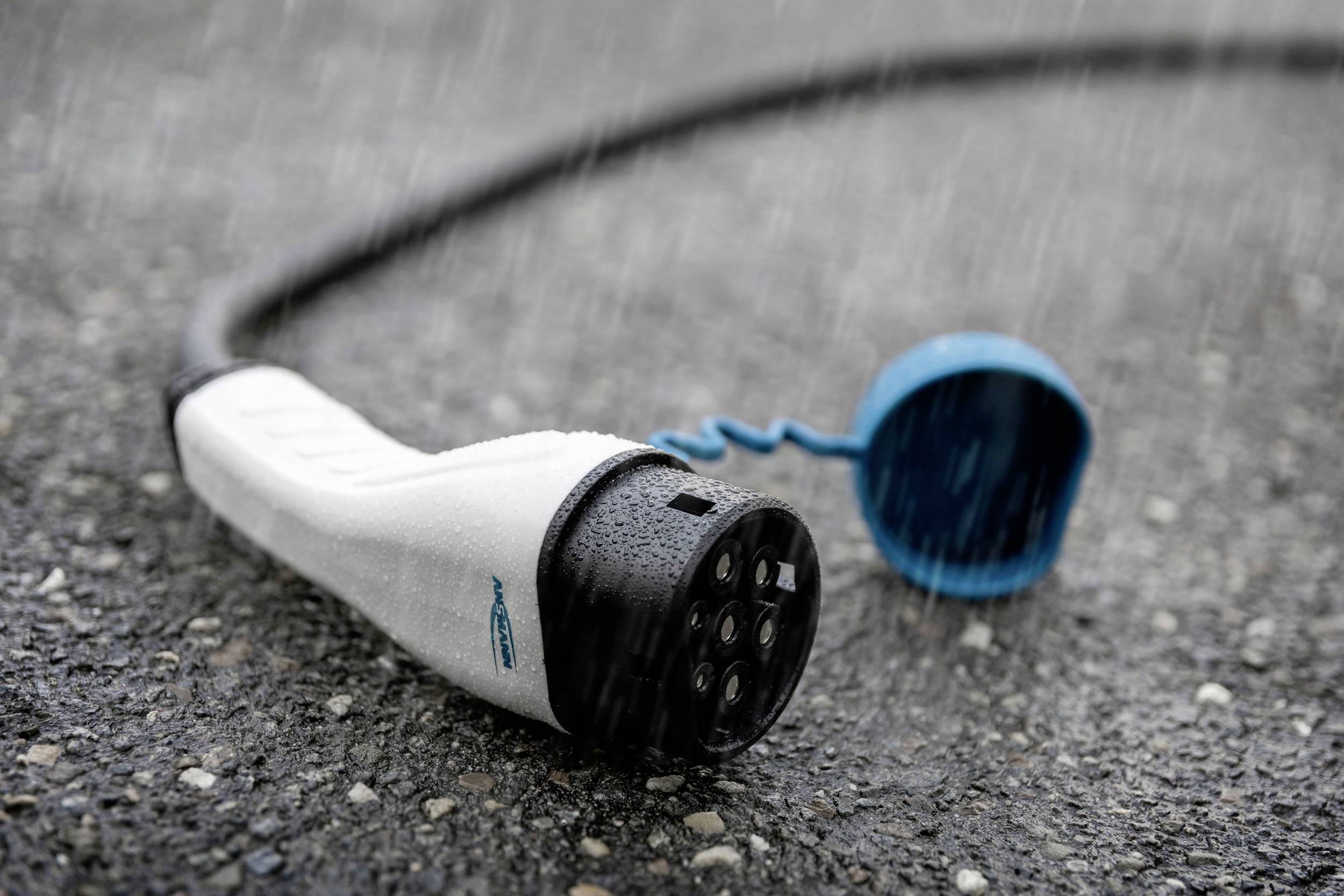 Ein Elektroauto-Ladekabel liegt auf nassem Asphalt im Regen. Der Stecker ist weiß mit einer blauen Schutzkappe.