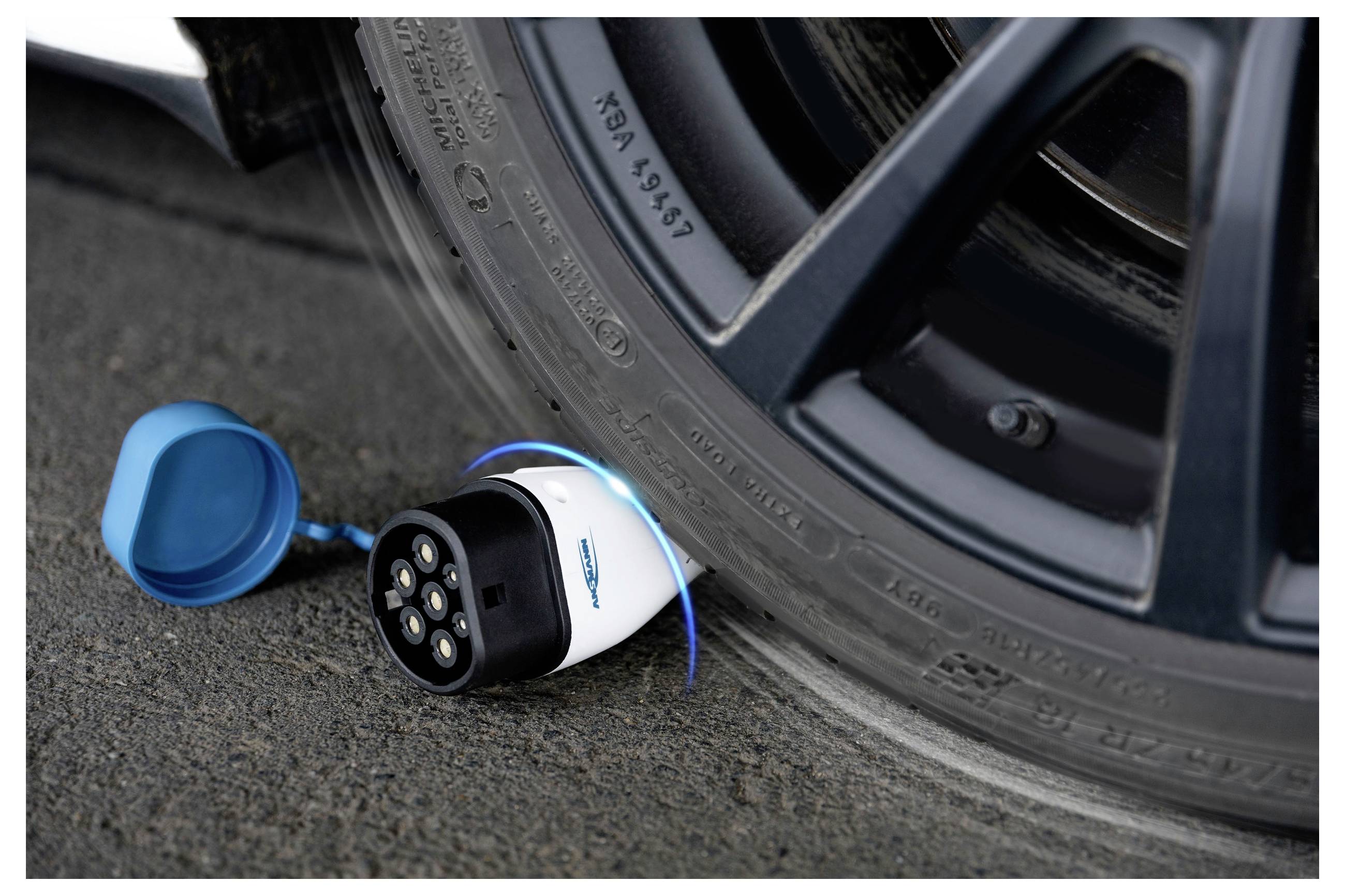 Ein elektrischer Ladestecker liegt ungenutzt auf dem Boden, eingeklemmt unter einem Autoreifen.