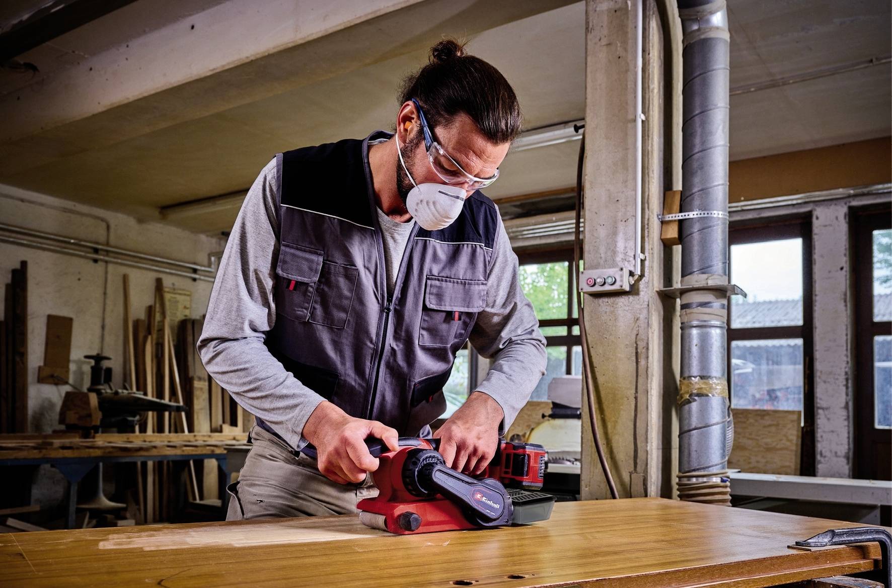 Eine Person schleift mit einer elektrischen Schleifmaschine Holz in einer Werkstatt. Sie trägt eine Schutzbrille und eine Atemmaske.