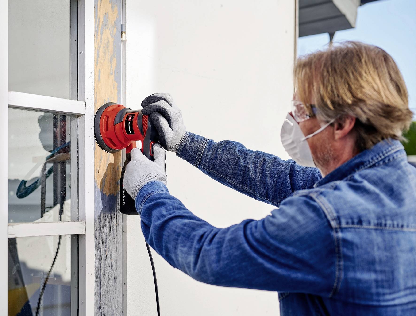 Ein Mann schleift den Holzrahmen eines Fensters mit einer elektrischen Schleifmaschine ab. Er trägt Schutzbrille und Maske.
