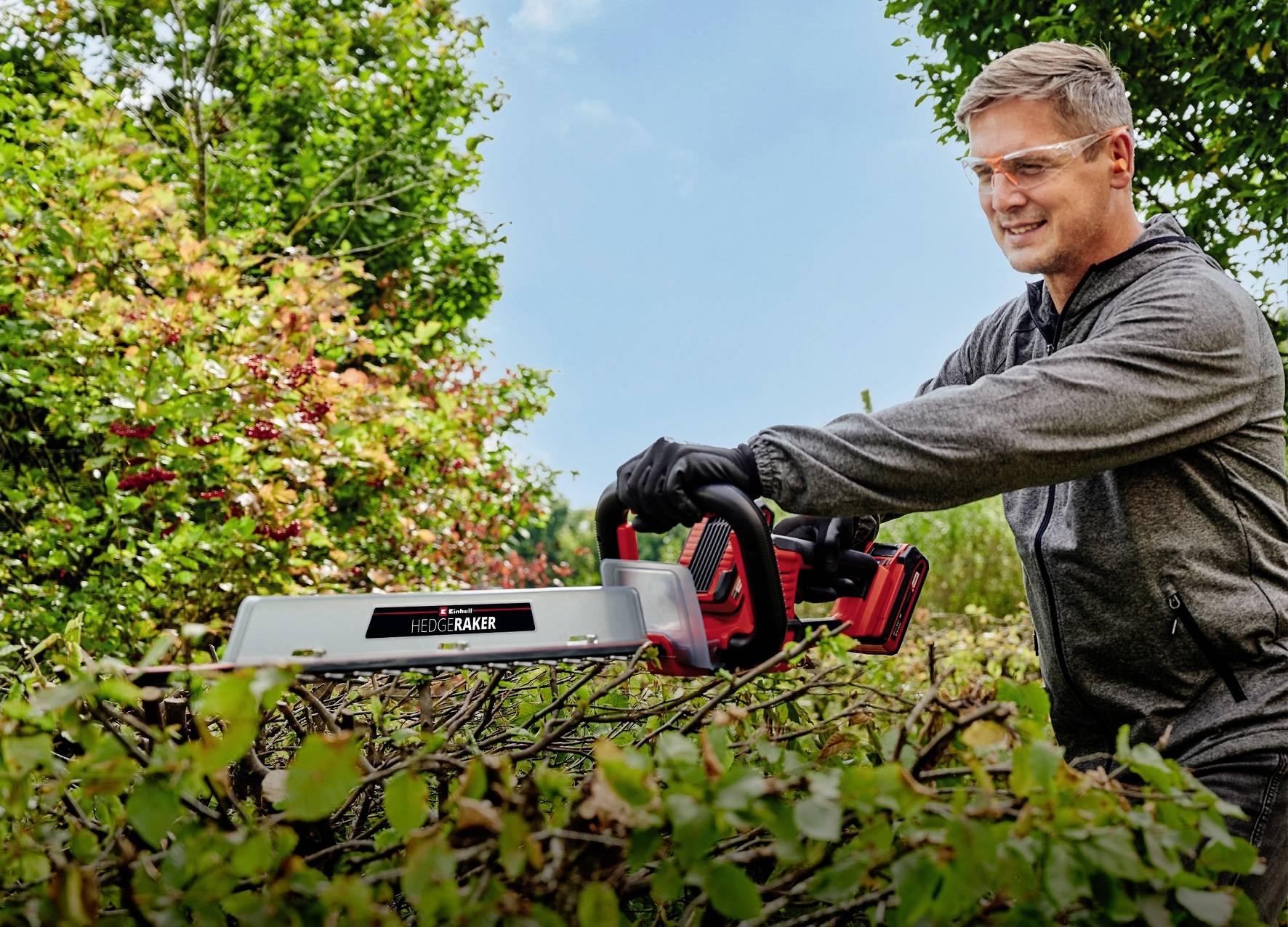 Ein Mann schneidet mit einer elektrischen Heckenschere Büsche im Garten. Er trägt Schutzbrille und Handschuhe, lächelt konzentriert.