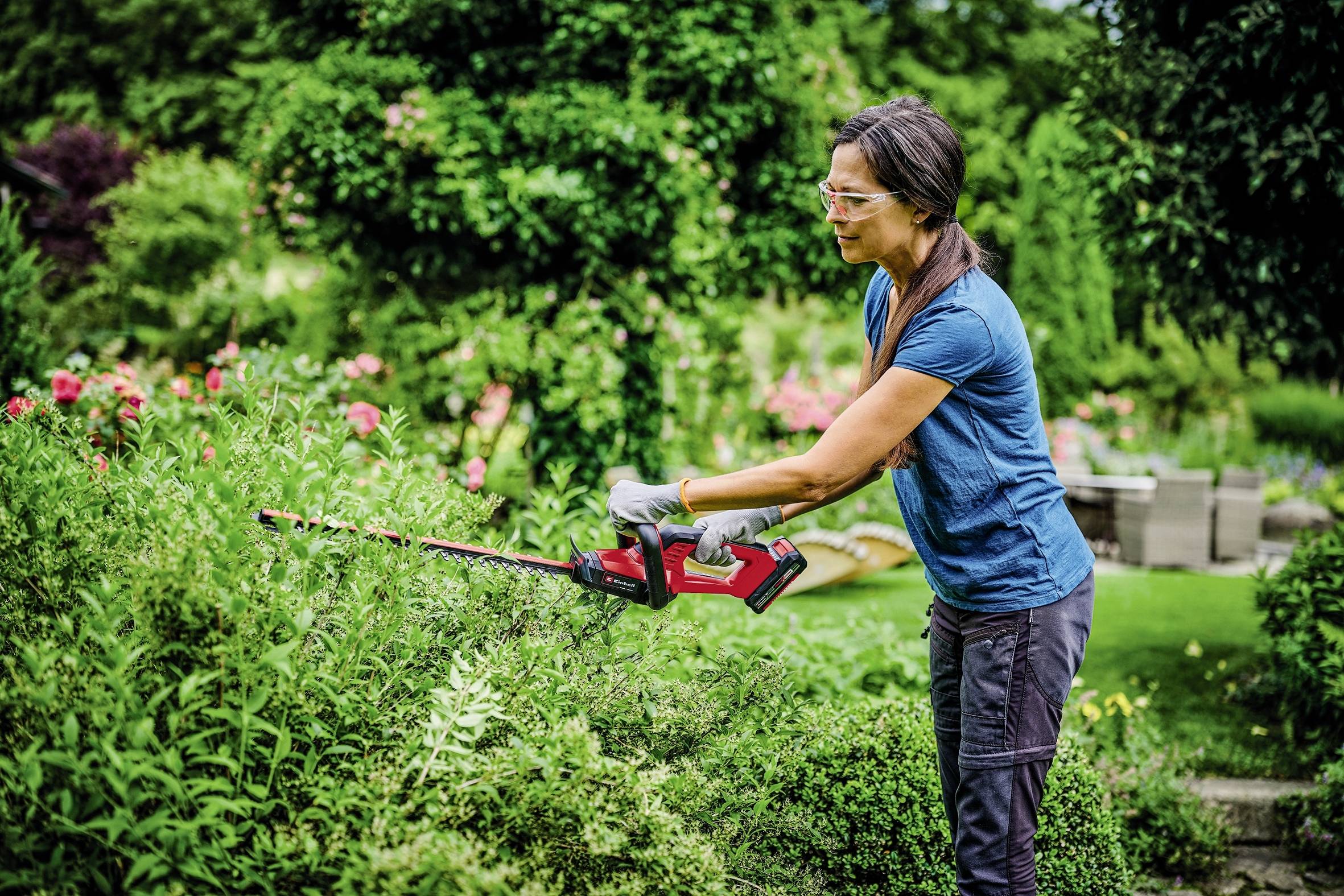 Frau schneidet Sträucher im Garten mit einer elektrischen Heckenschere. Im Hintergrund sind Büsche und Sitzmöbel zu sehen.