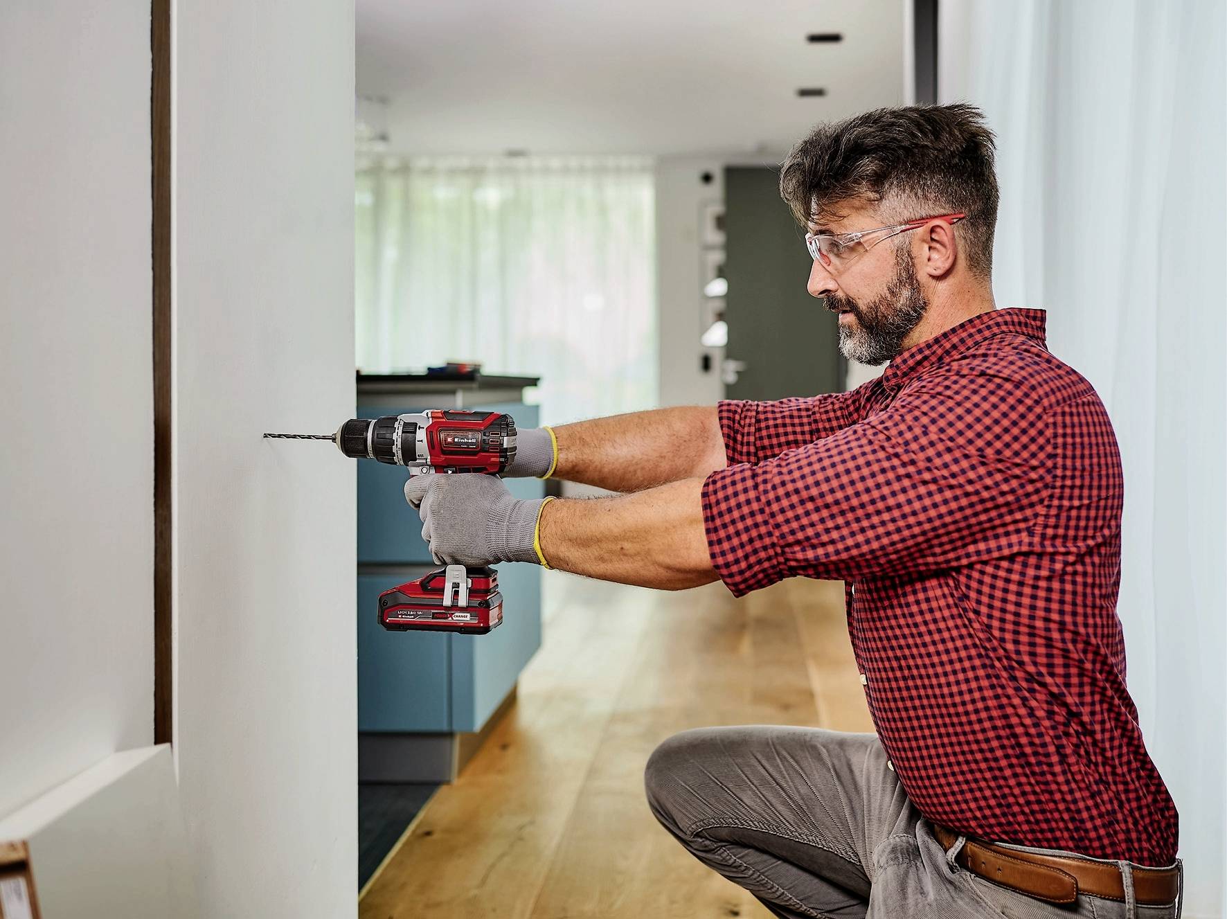 Ein Mann in einem karierten Hemd benutzt einen Bohrer, um in einer modernen Wohnung zu arbeiten. Er trägt Schutzbrille und Handschuhe.