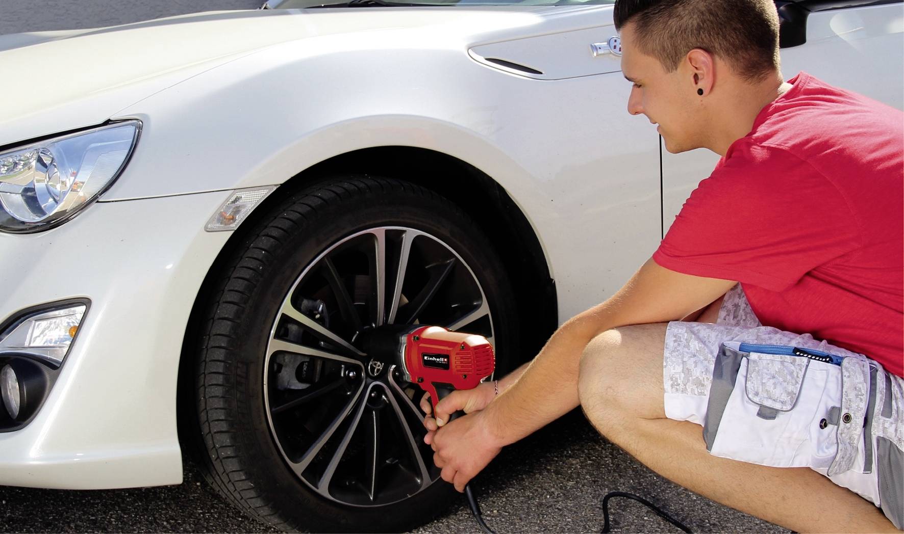 Ein Mann in rotem T-Shirt und kurzer Hose wechselt mit einem Schraubenschlüssel einen Autoreifen bei einem weißen Auto.