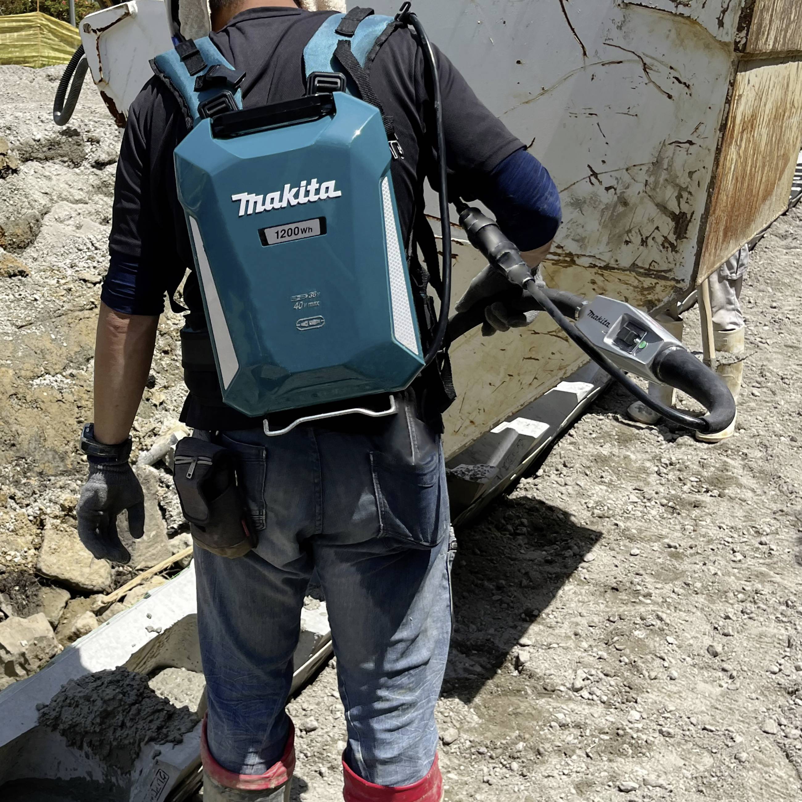 Eine Person auf einer Baustelle trägt einen großen Makita-Rucksack und bewegt ein schweres, fahrbares Gerät über unebenes Gelände.