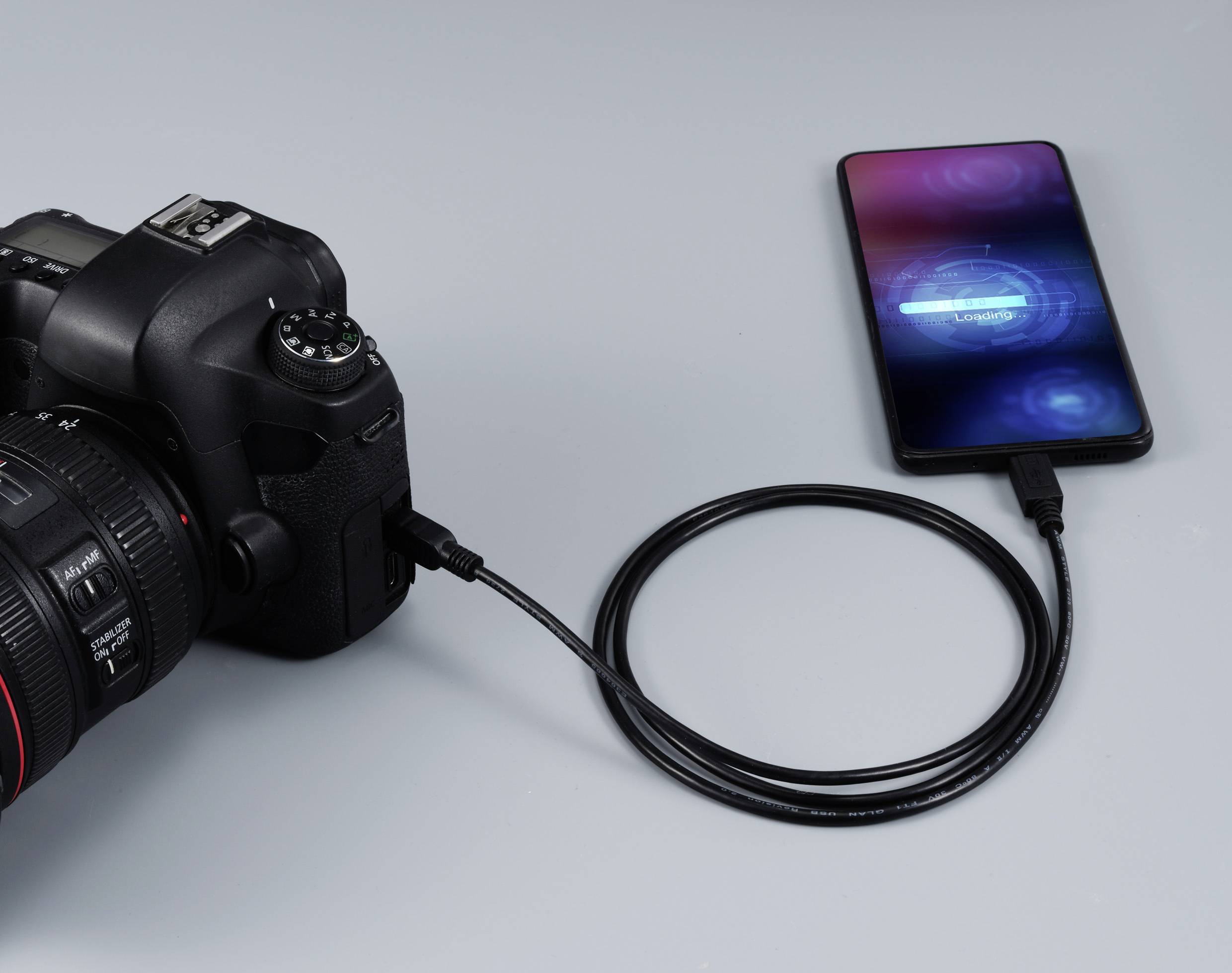 Kamera verbunden mit einem Smartphone durch ein USB-Kabel. Auf dem Smartphone-Bildschirm steht 'Ladegerät wird verwendet'.