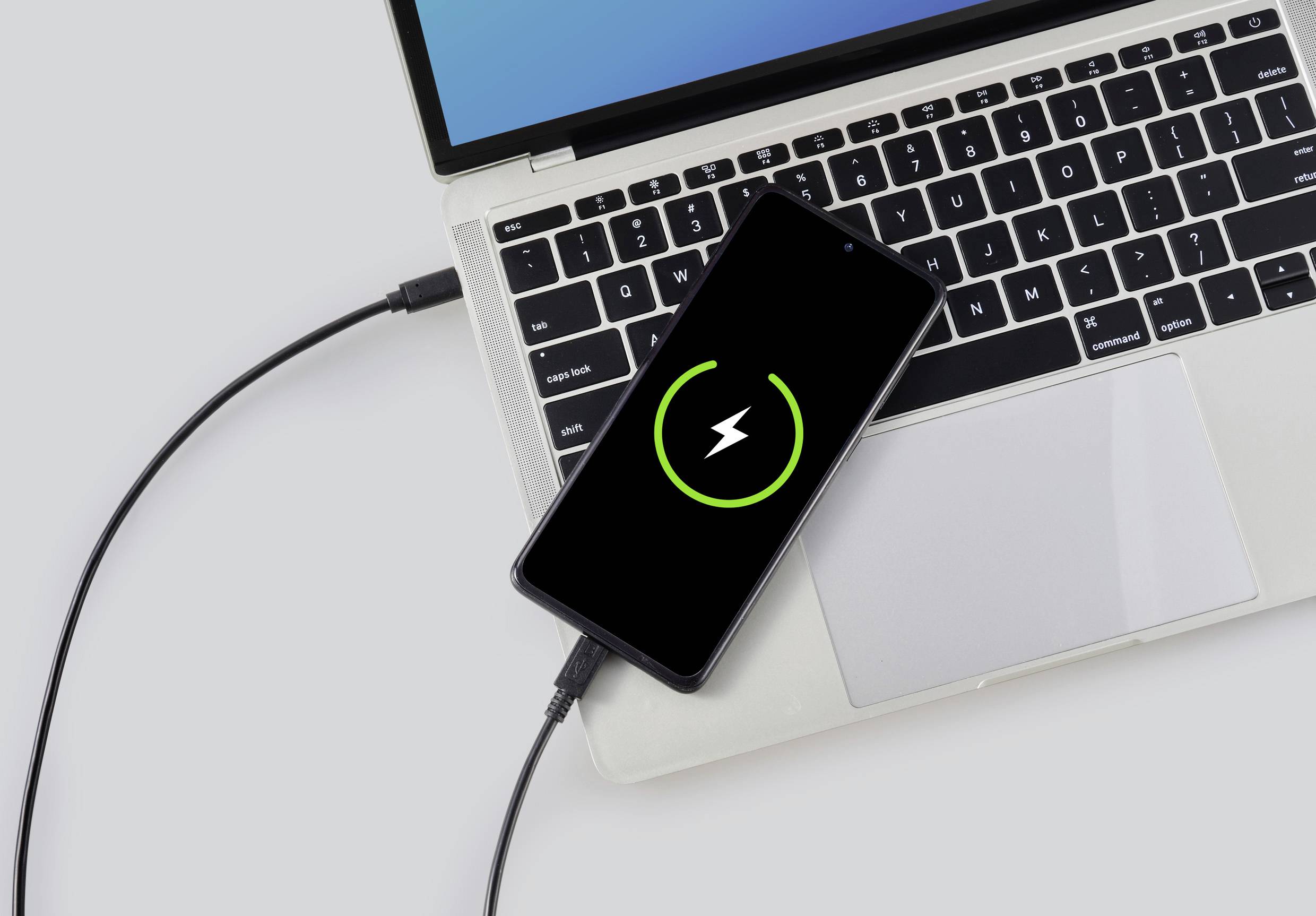 Ein Smartphone liegt auf der Tastatur eines Laptops und wird über ein Kabel aufgeladen, der Ladebildschirm zeigt einen grünen Kreis mit Blitzsymbol.
