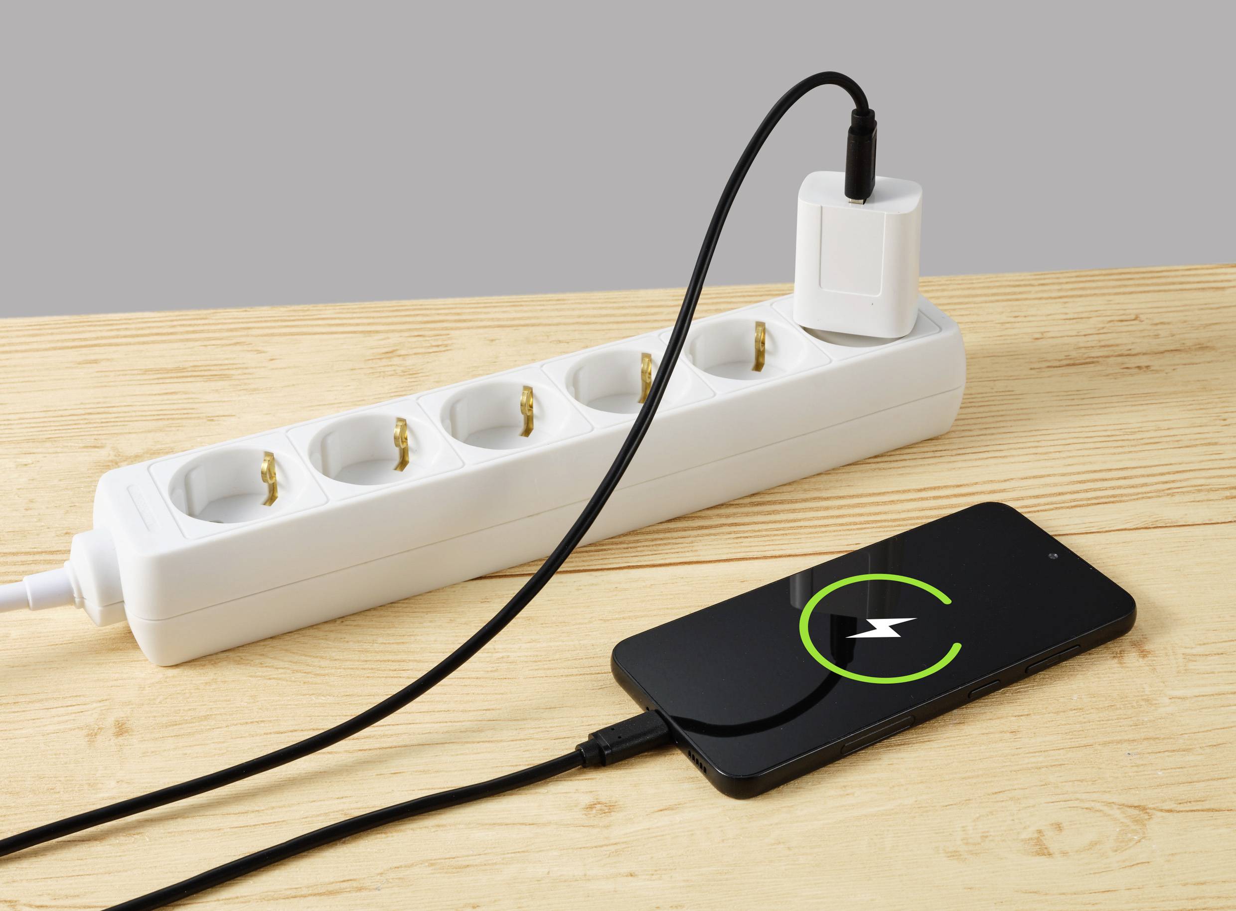 Ein Smartphone wird über ein Ladekabel und einen Adapter, der in eine Steckdosenleiste eingesteckt ist, auf einem Holztisch geladen.