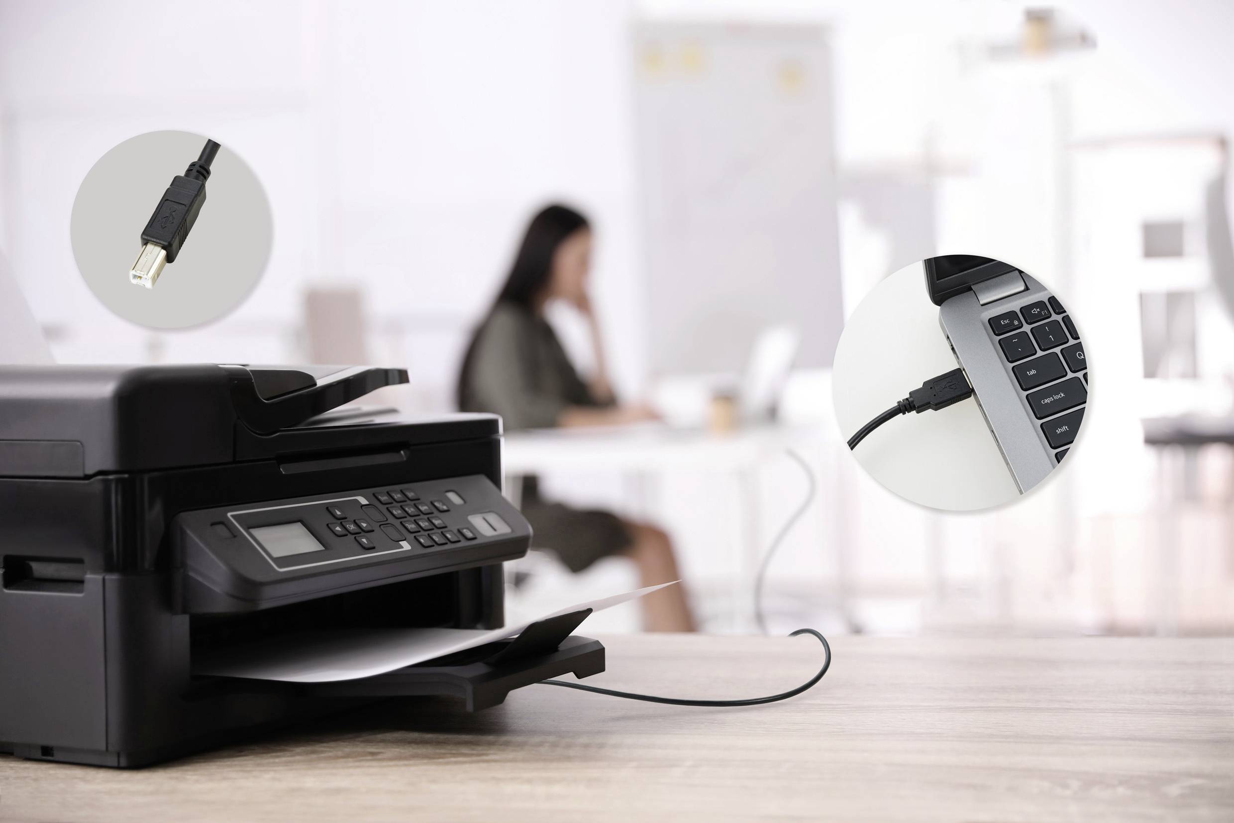 Ein Drucker auf einem Schreibtisch ist über ein Kabel mit einem Laptop verbunden. Im Hintergrund eine Person unscharf sitzend an einem Tisch.