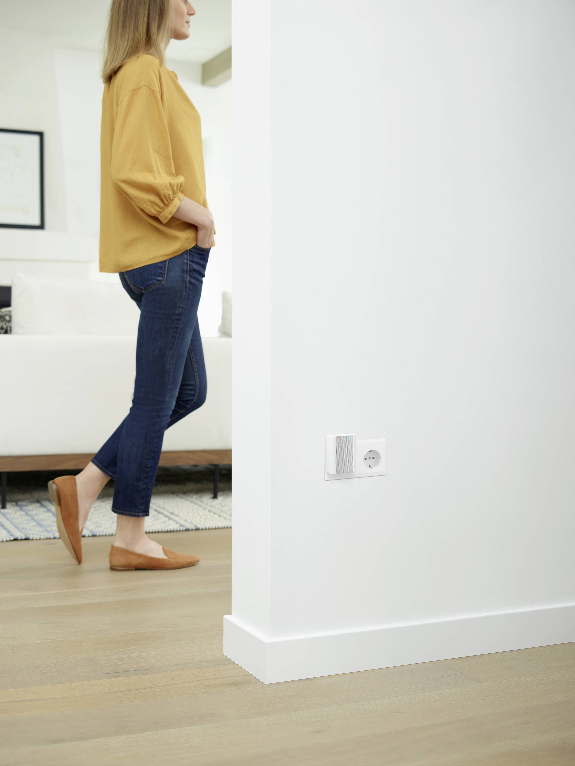 Person in gelbem Pullover und Jeans steht in modernem Wohnzimmer; im Vordergrund ist eine Steckdose mit einem kleinen Gerät daran sichtbar.